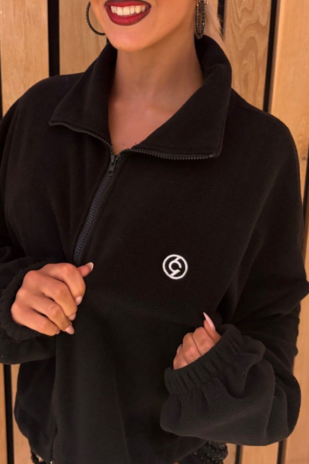 A woman wearing a black half-zip fleece pullover with a small white circular logo on the left chest, standing in front of a vertical wood slat wall.