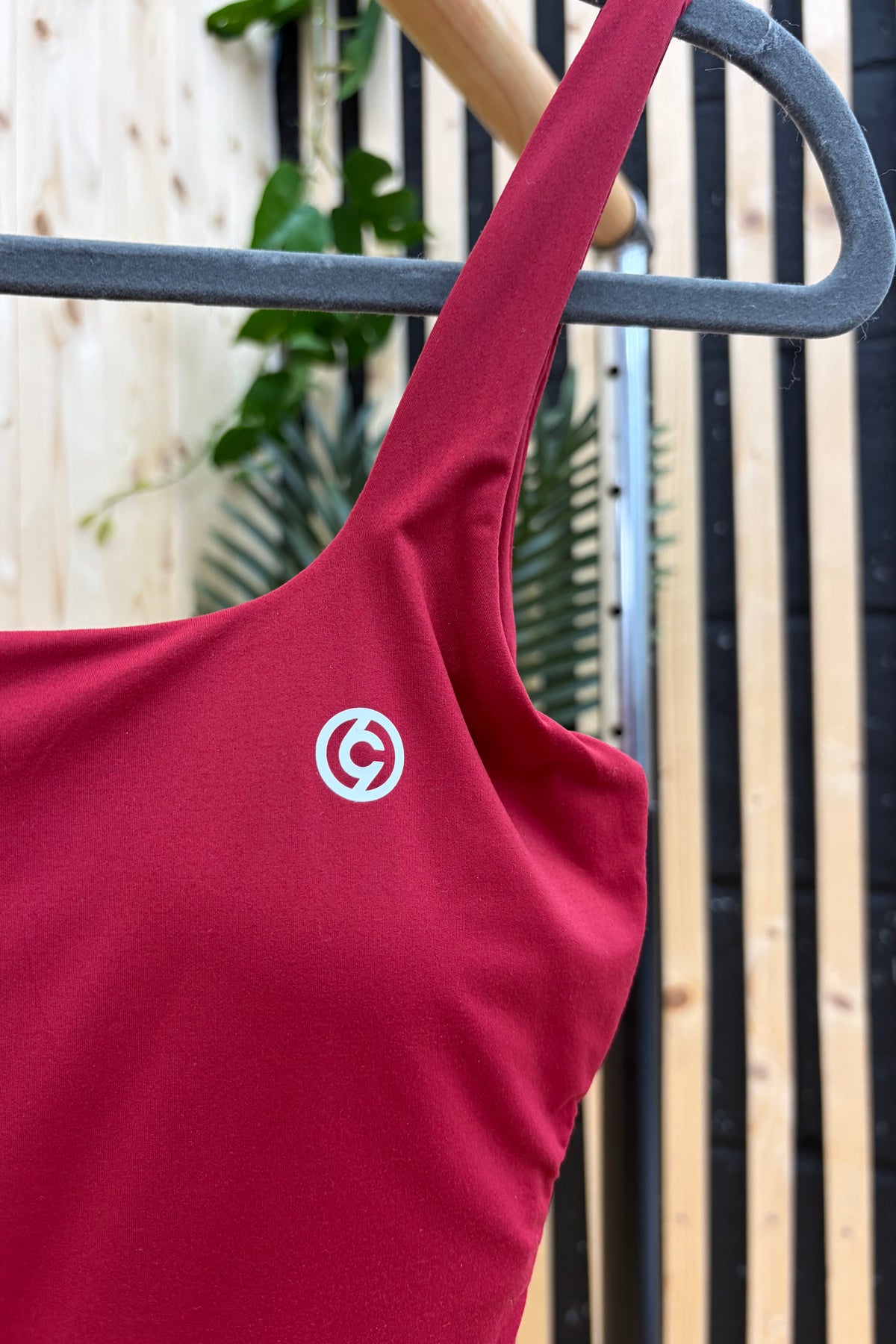 A close-up of a red one-piece swimsuit or tank top hanging on a gray hanger, featuring a small white circular logo with a stylized 'G' on the chest. The background shows a wooden fence and green foliage.