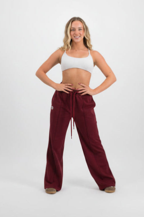 A woman modeling a pair of wide-leg burgundy sweatpants with a white sports bra, standing against a light gray studio background.