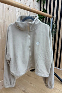 A light beige fleece half-zip pullover jacket hanging on a wooden hanger from a clothing rack, set against a background of light wood paneling and a black slatted screen.