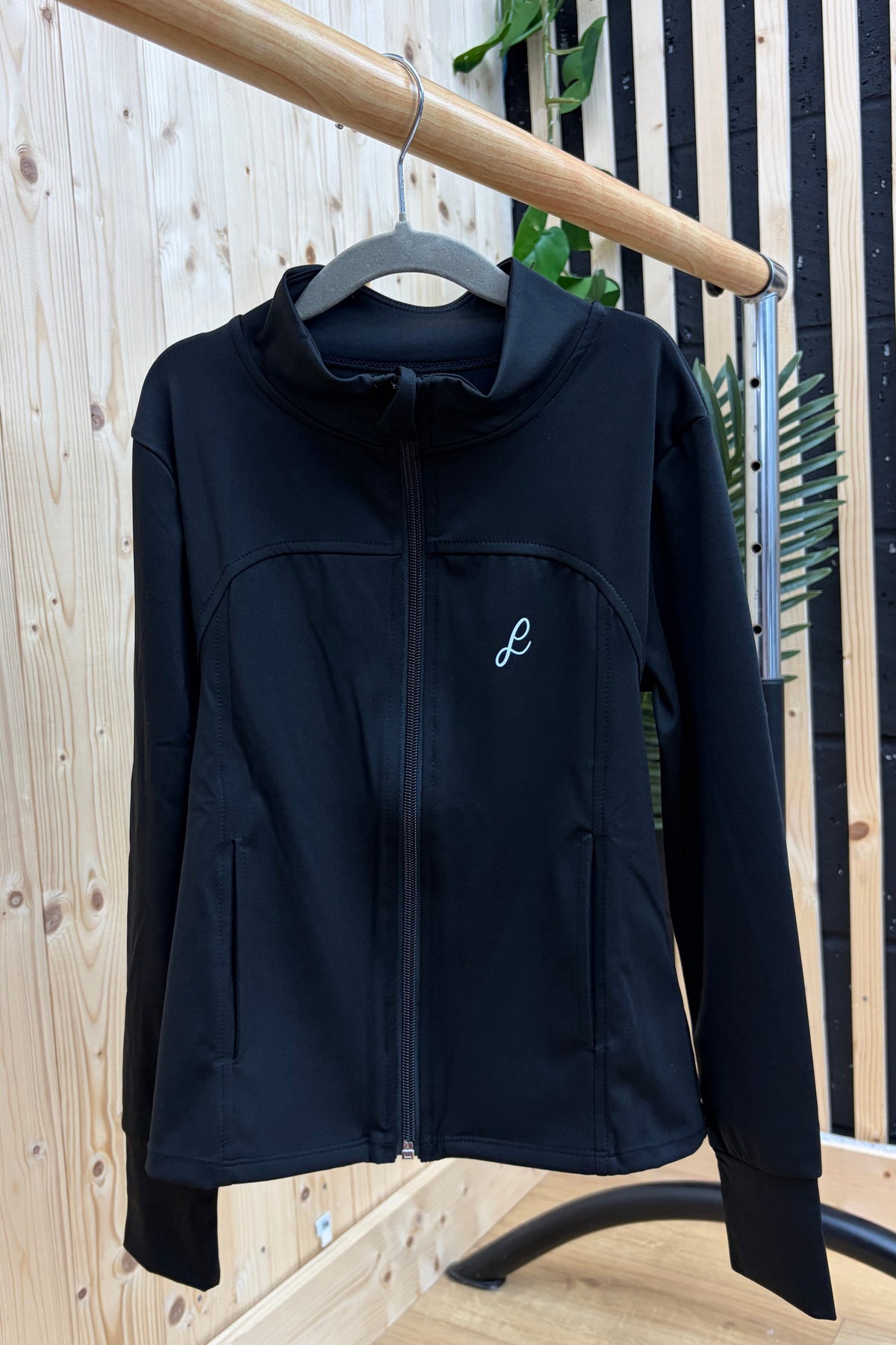 A black hooded zip-up jacket hanging on a gray hanger from a wooden clothing rack, set against a background of light wood paneling and a black-and-wood slatted wall.