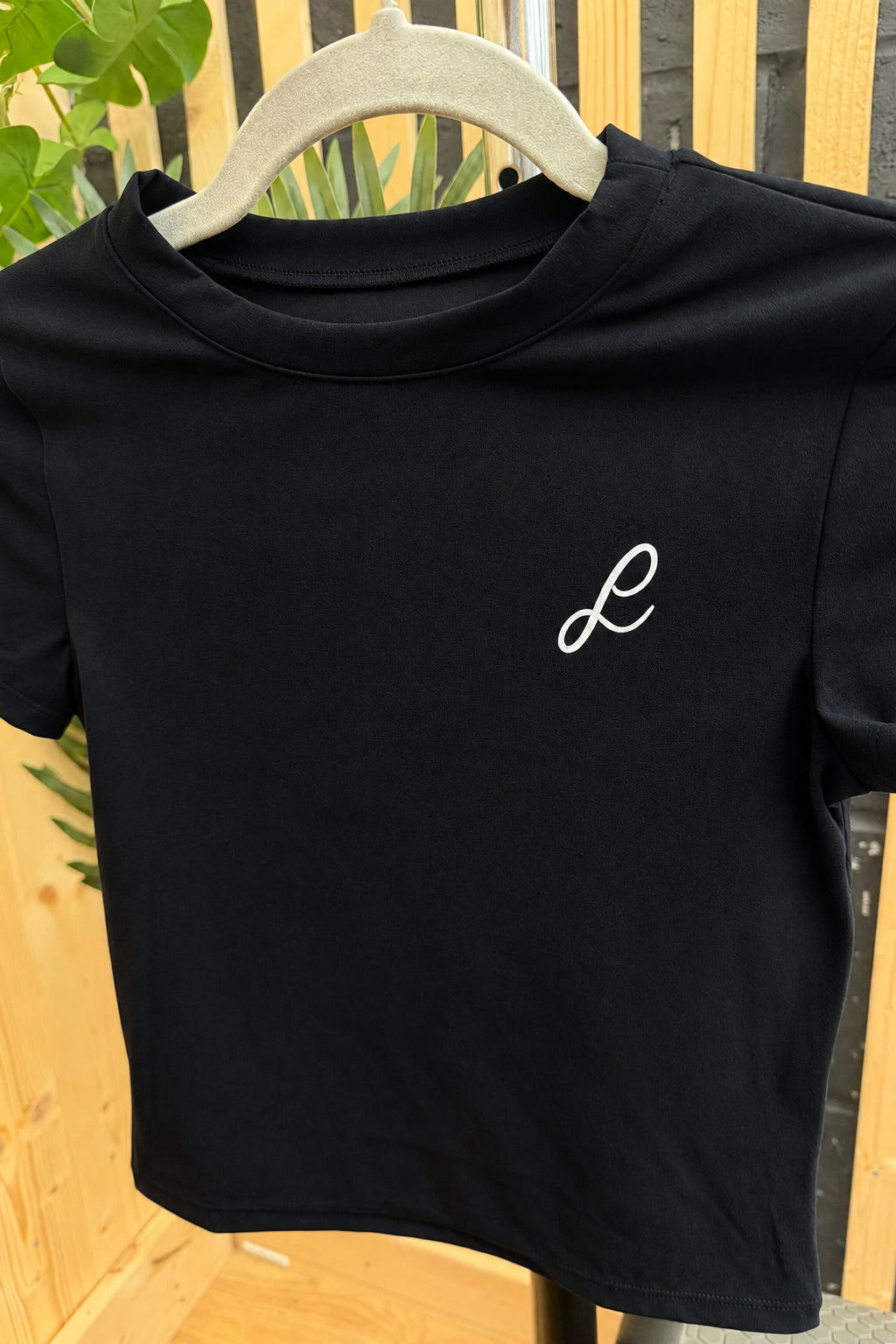 A black long-sleeve athletic zip-up jacket with a white embroidered script logo on the left chest, displayed on a hanger against a wooden slat background.