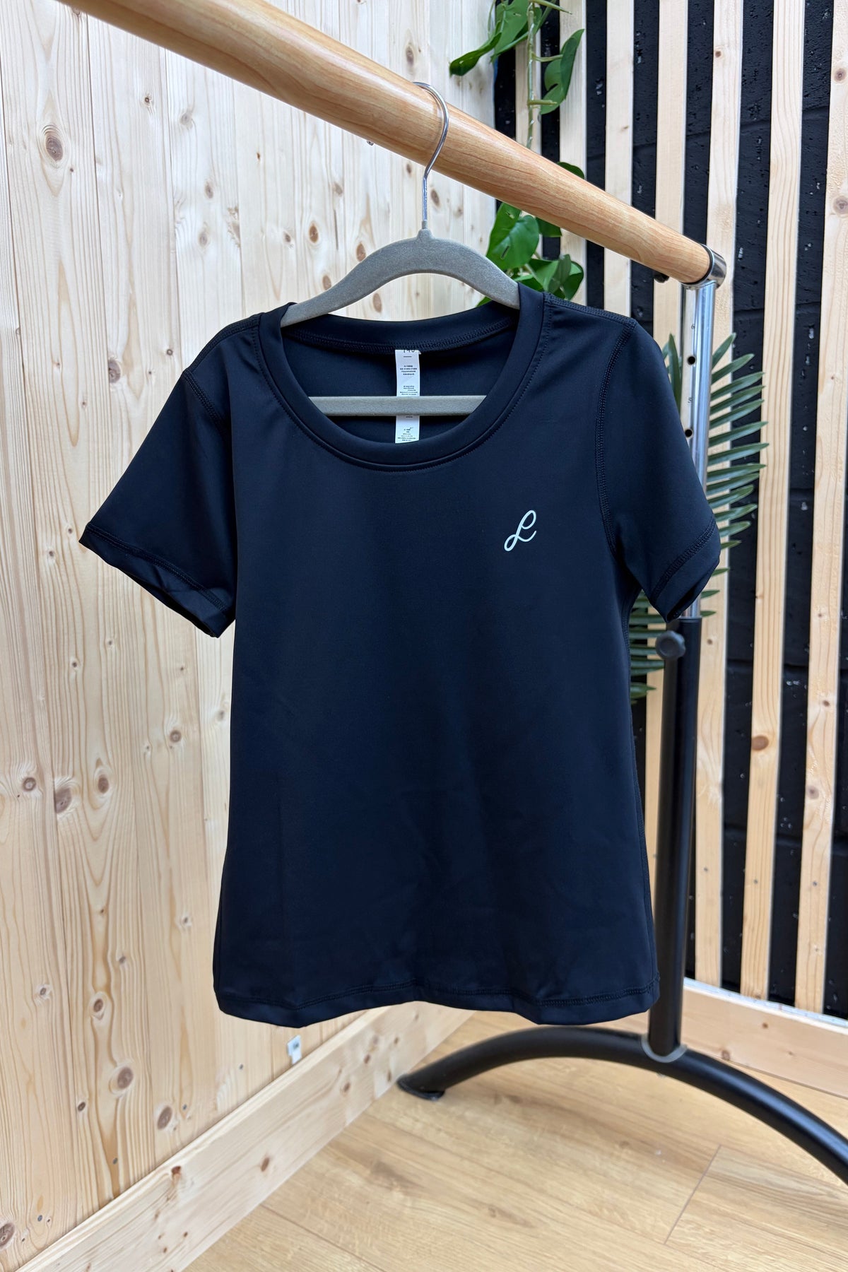 A black short-sleeve crewneck t-shirt with a small white script logo on the left chest, hanging on a gray hanger from a wooden clothing rack in front of a light wood paneled wall.