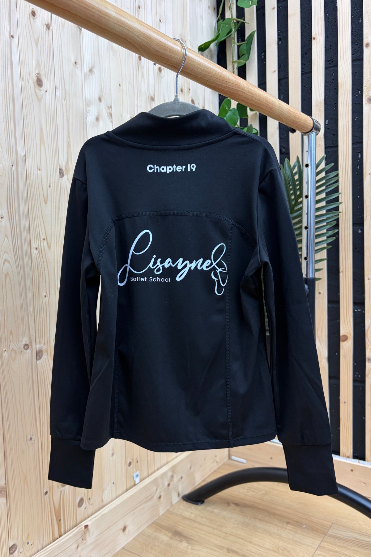 A black long-sleeve athletic jacket with white custom text and logo graphics on the back, hanging on a wooden clothing rack against a light wood-paneled wall.