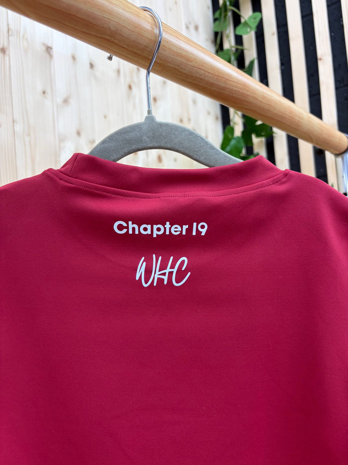 A close-up of a dark red crewneck sweatshirt hanging on a gray hanger from a wooden clothes rack. The back of the sweatshirt features white printed text reading 'Chapter 19' and 'WHC'.