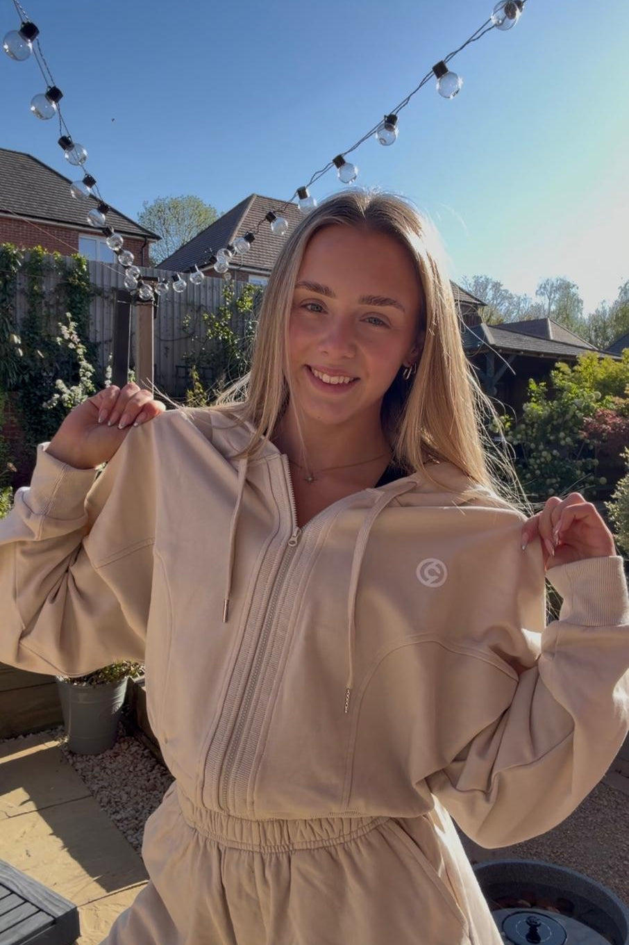 Woman wearing a beige tracksuit with a logo, standing outdoors with string lights and houses in the background.