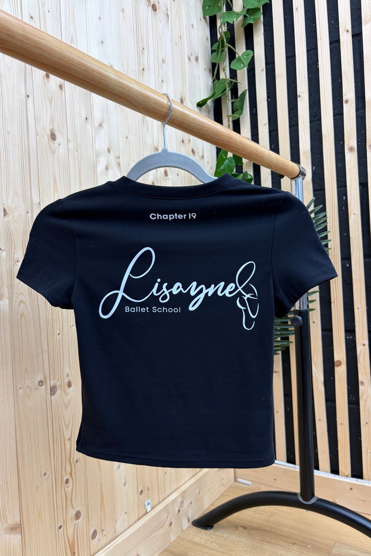 A navy blue short-sleeve t-shirt with custom white text reading 'Chapter 19 Lisaynel Ballet School' on the back, hanging on a gray hanger from a wooden clothing rack in a styled indoor setting.
