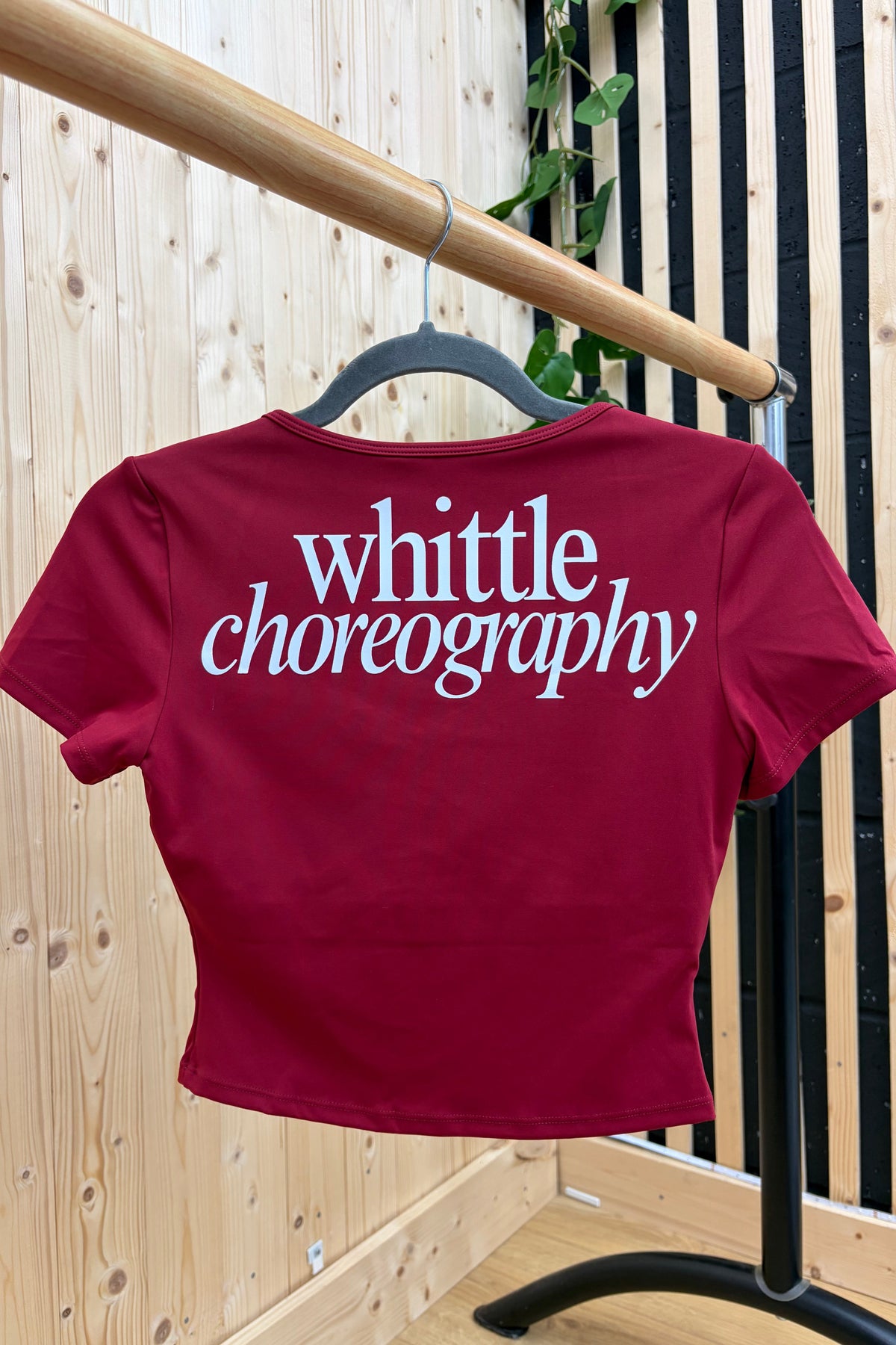 A maroon short-sleeve crop top with 'whittle choreography' printed in white script across the back, hanging on a black hanger from a wooden clothing rack.