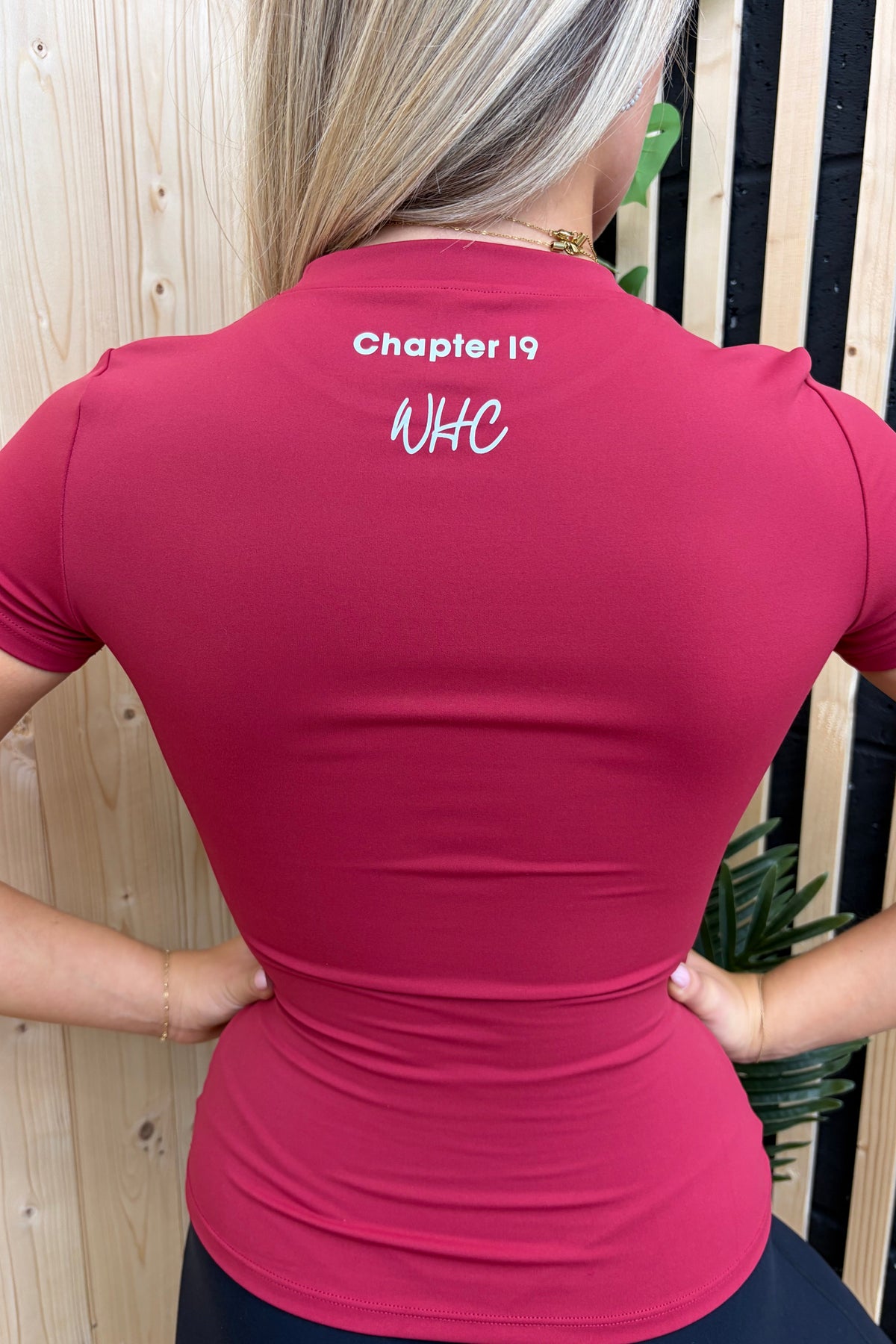 A woman viewed from behind wearing a fitted short-sleeve athletic top in a deep pink/magenta color. The back of the shirt features white printed text reading 'Chapter 19' and 'WKC'.
