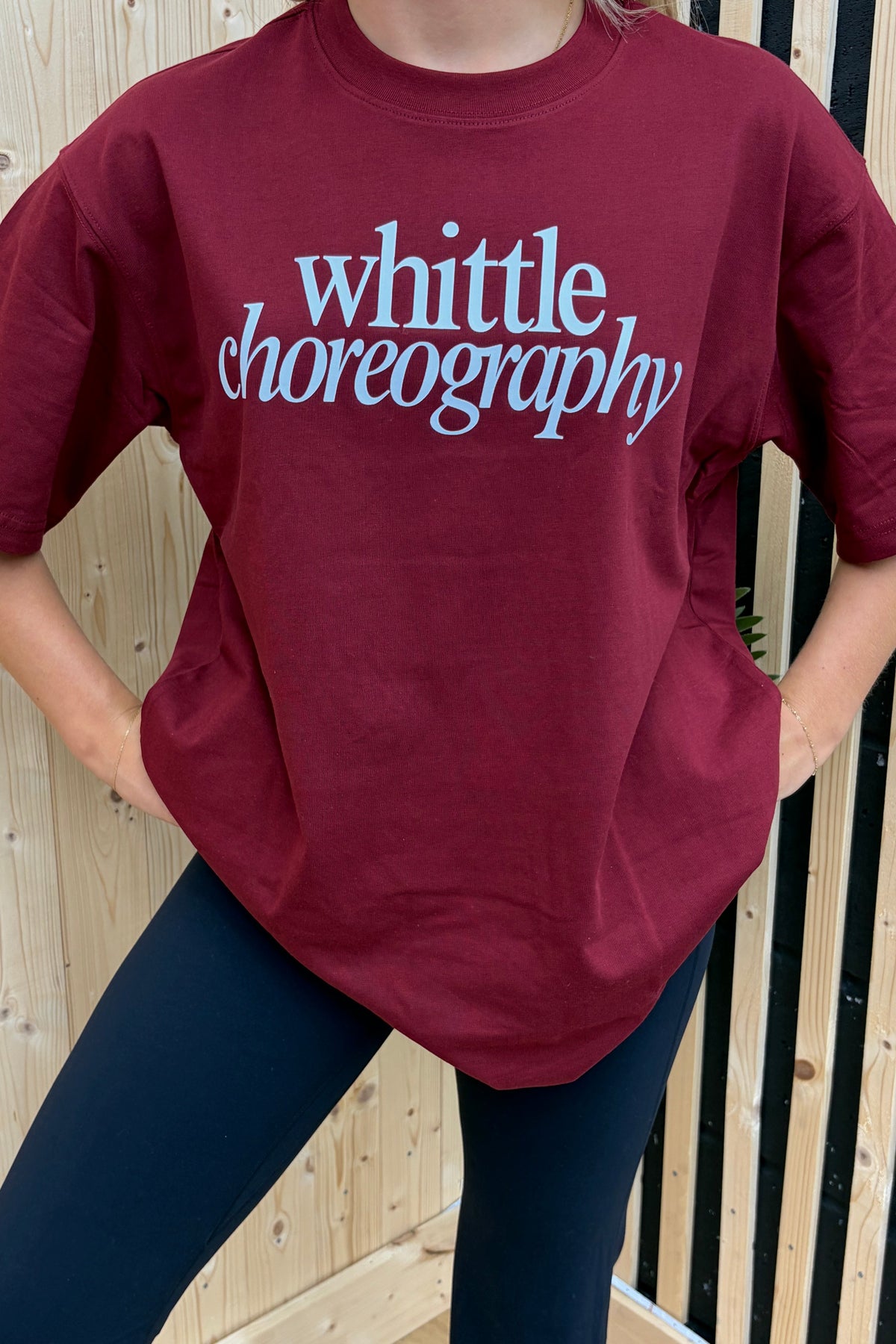 A person wearing an oversized burgundy t-shirt with 'whittle choreography' printed in light blue script across the chest, paired with dark leggings, standing against a light wood slat wall.