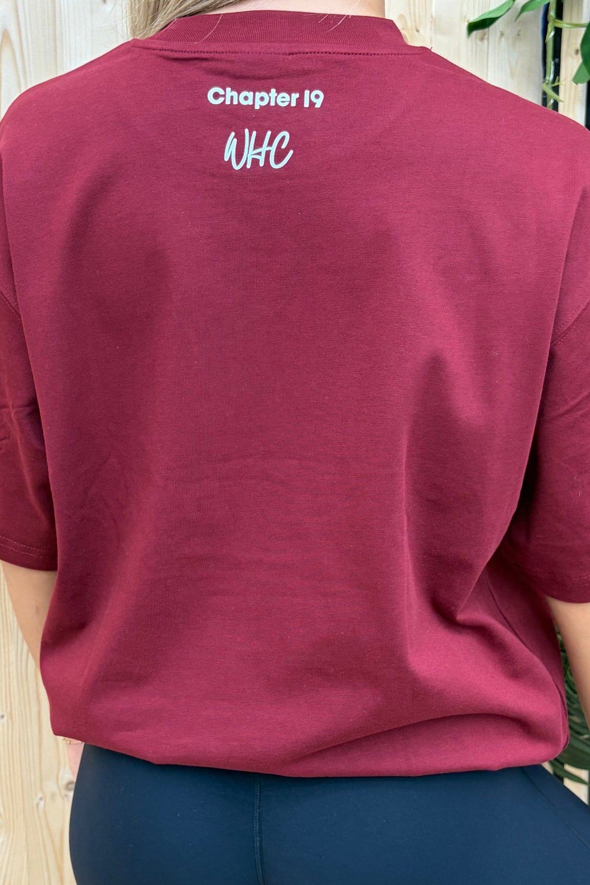 A woman shown from the back wearing an oversized short-sleeve t-shirt in burgundy with white text reading 'Chapter 19' and 'NHC' on the upper back, paired with dark leggings, set against a light wood slat wall background.