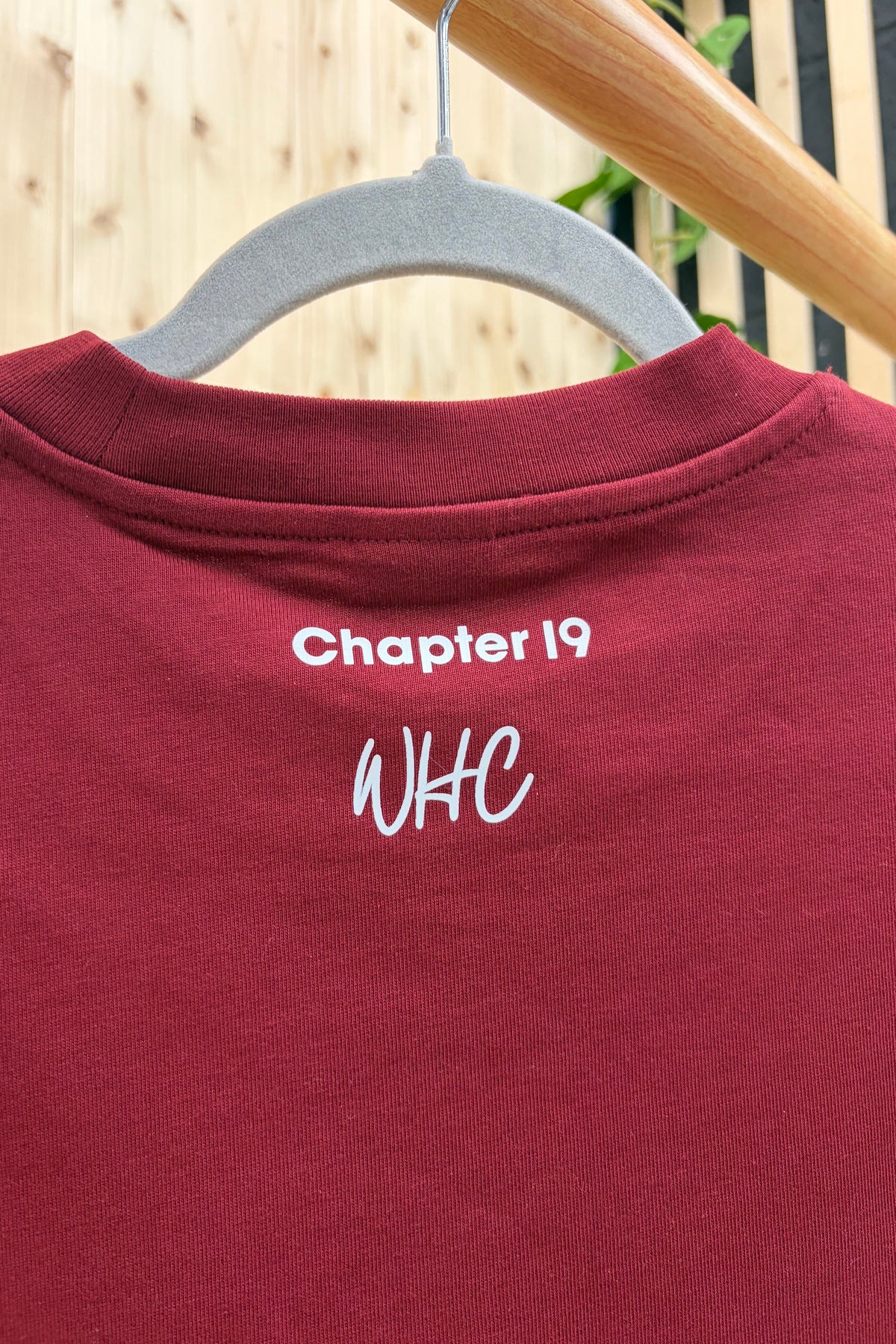 A close-up of a maroon crewneck t-shirt hanging on a gray plastic hanger. The shirt features white embroidered text on the back collar reading 'Chapter 19' and 'WHC'.