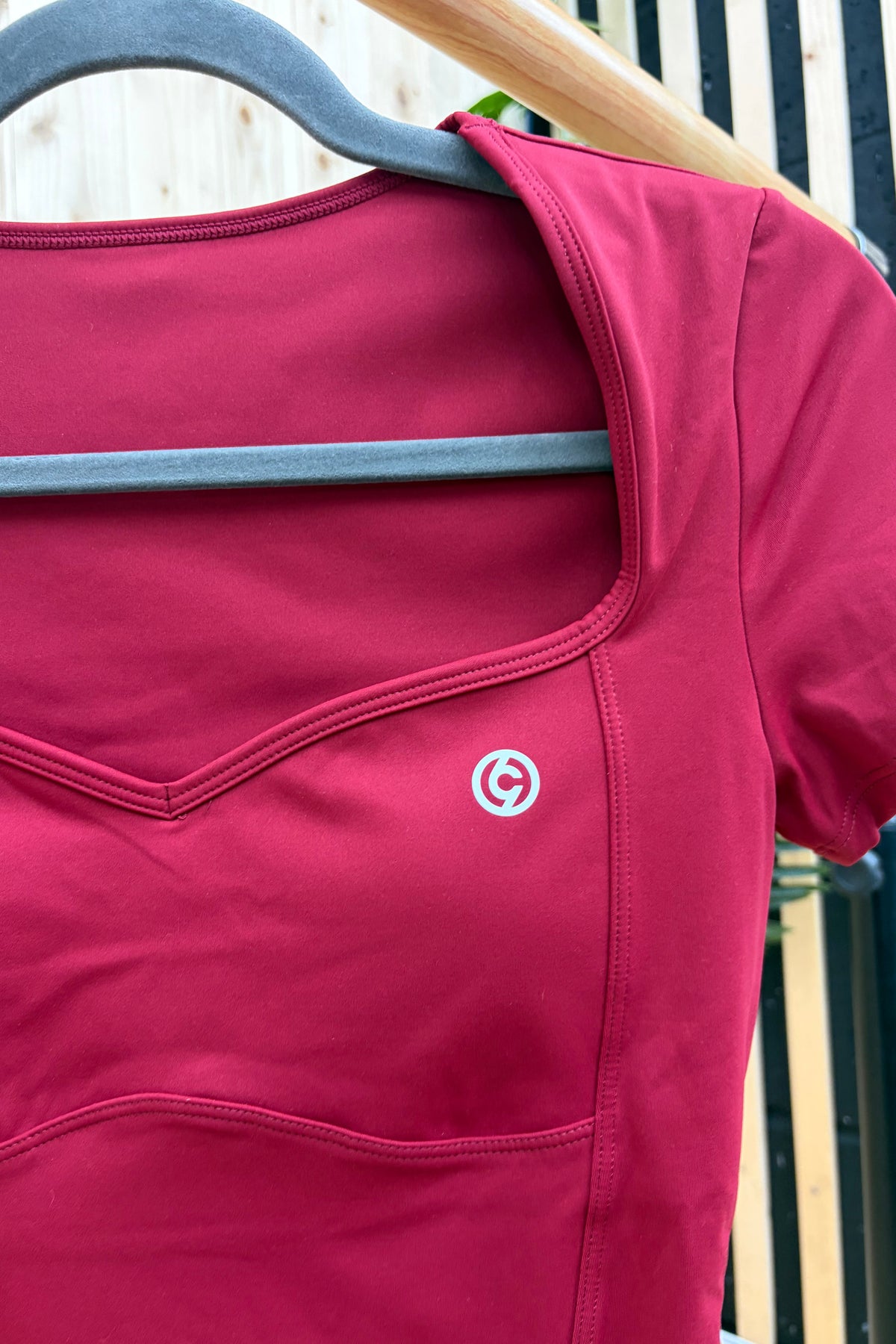 A close-up of a short-sleeve athletic top in deep magenta pink, hanging on a gray hanger. The top features a scoop neckline, curved seam details, and a small circular white and red logo on the chest.