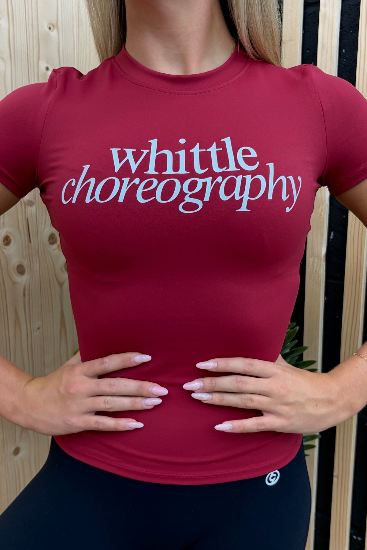 A woman wearing a fitted short-sleeve athletic top in deep red with the words 'whittle choreography' printed across the chest in light blue, posed with hands on hips against a light wood slat wall.