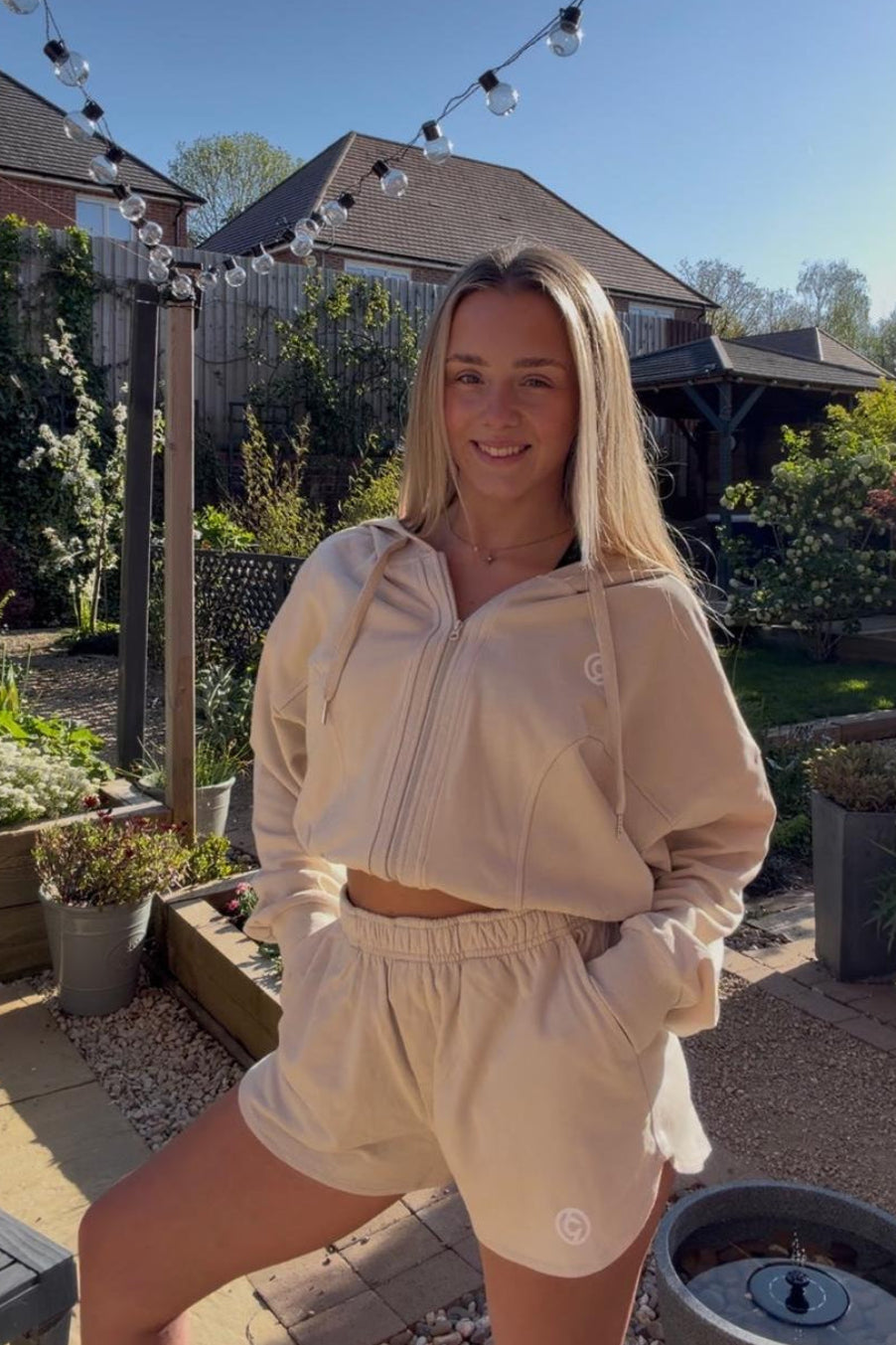 Woman in a beige tracksuit standing in a garden with string lights above.