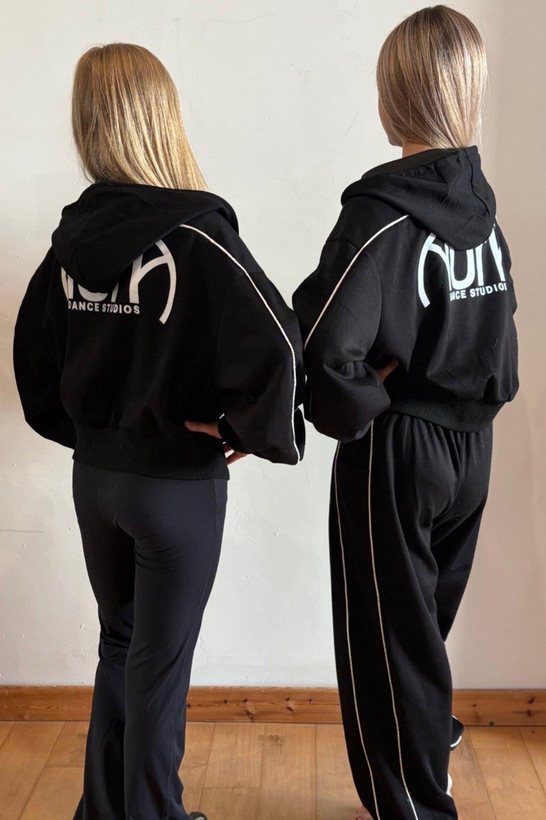 Two women standing from behind wearing matching black tracksuits with white side stripes and a white graphic logo on the back, in an indoor room with a wooden floor and off-white wall.