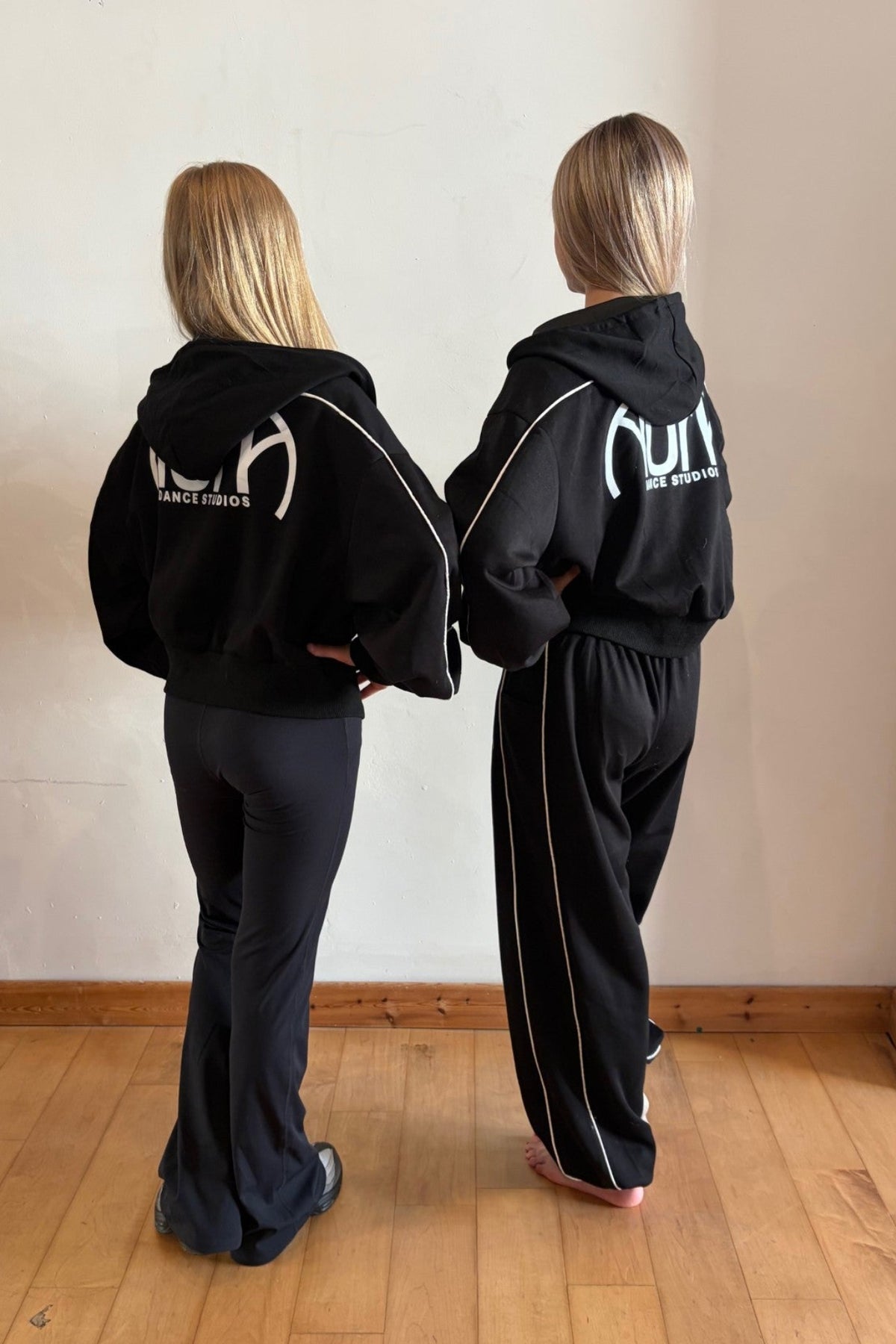 Two women standing from behind wearing matching black tracksuits with white side stripes and a white graphic logo on the back, in an indoor room with a wooden floor and off-white wall.