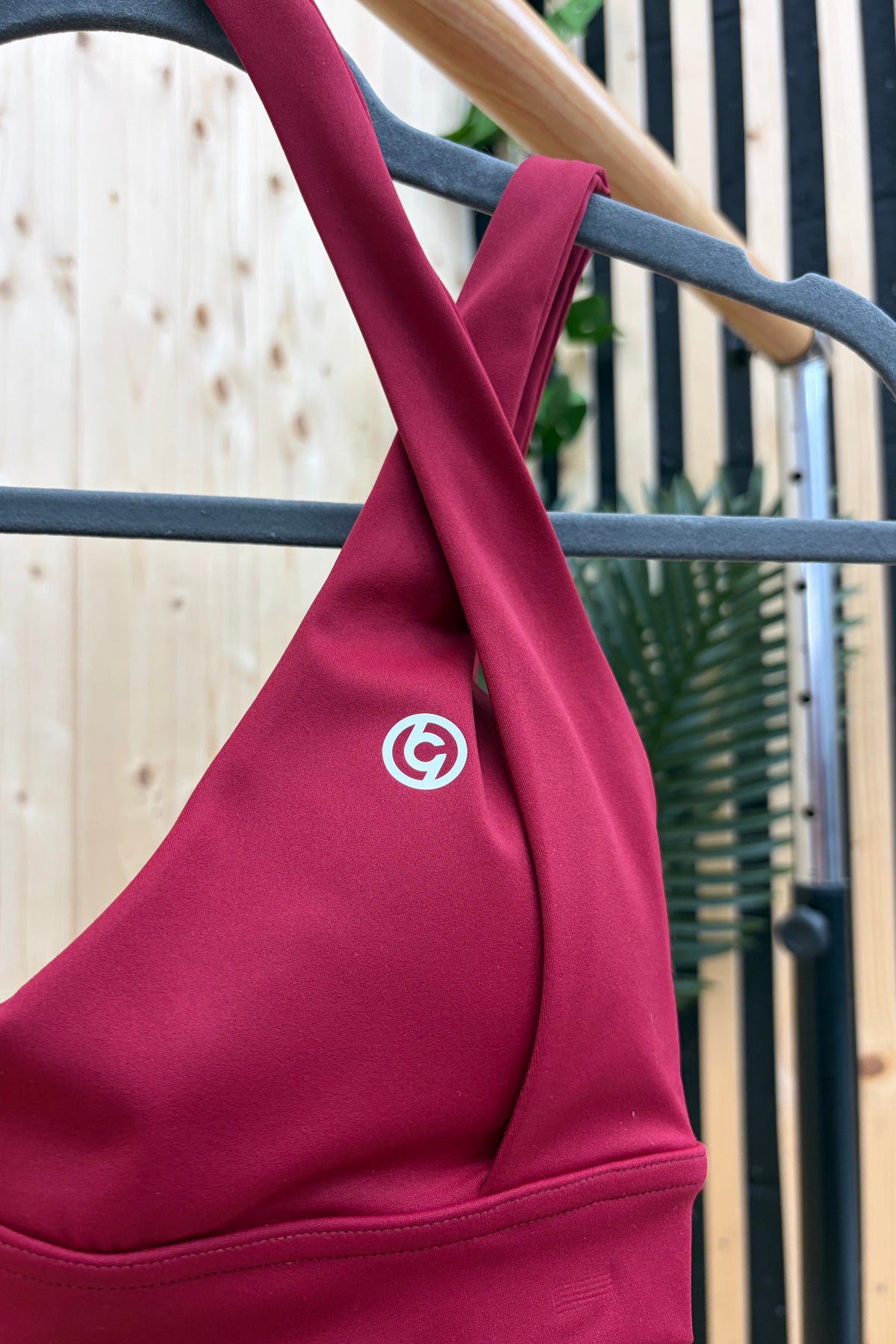 A close-up of a dark red sports bra hanging on a gray hanger, featuring a white circular logo on the chest and a crossover strap design, set against a background of a wooden slatted wall and green leaves.