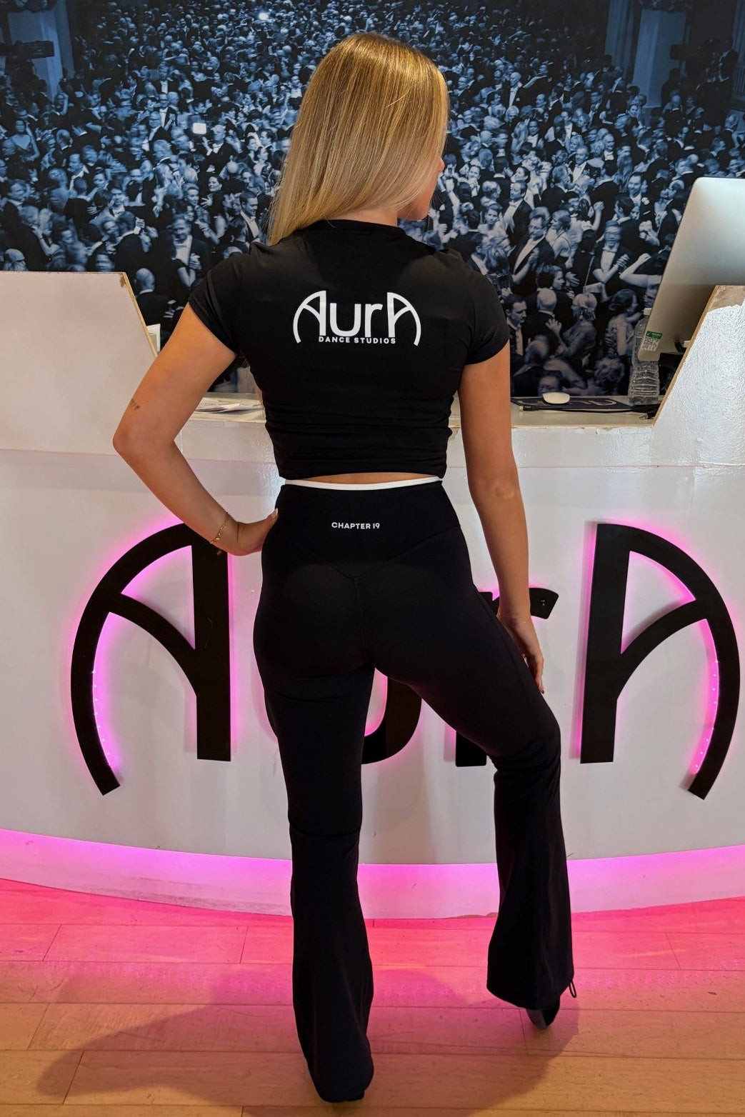 A woman stands with her back to the camera wearing a black crop top and black flared pants with the brand name 'Aura' printed on the back, posed in front of a white stage-like backdrop with pink lighting and a large audience photo in the background.