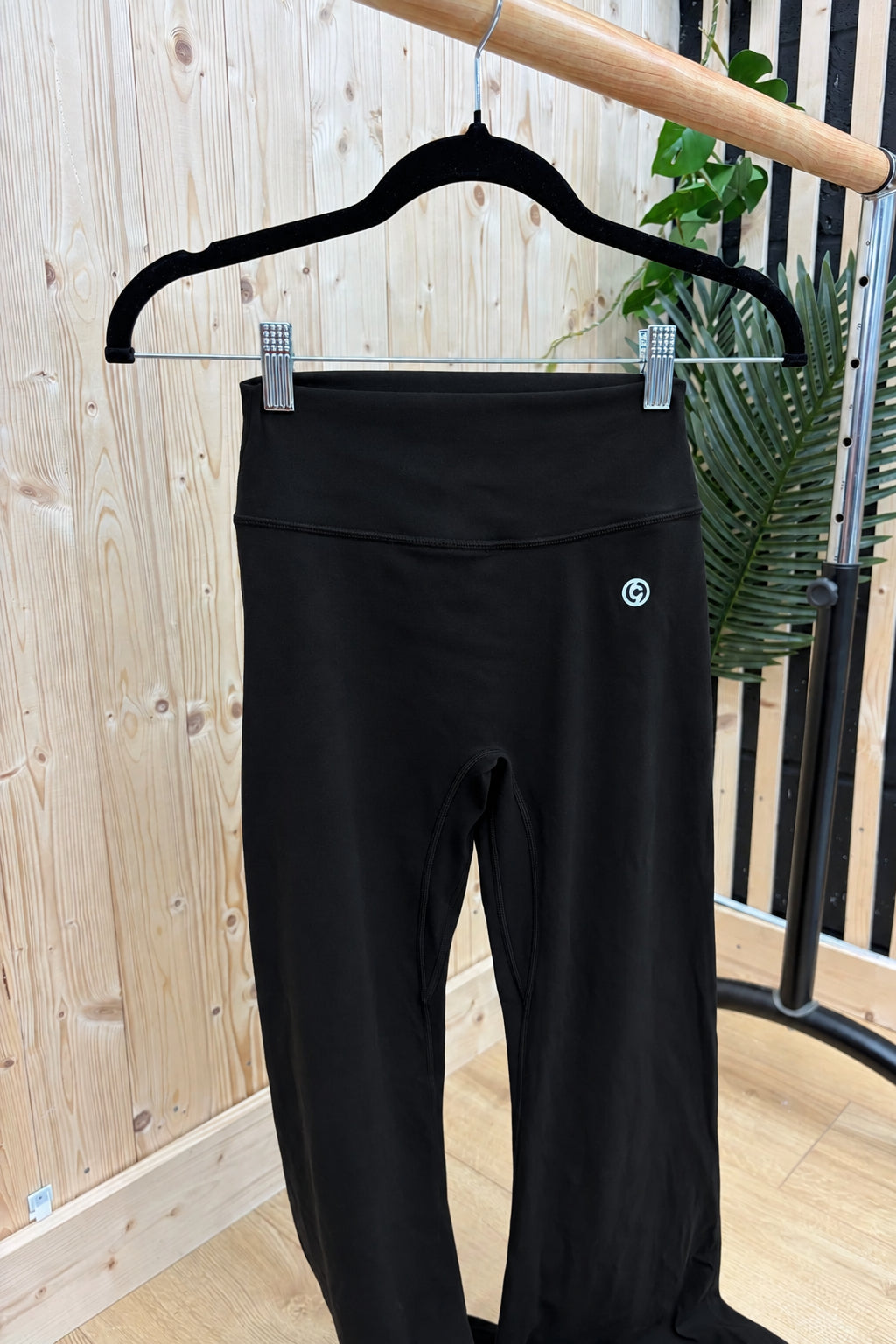 Black high-waisted leggings hanging on a black hanger from a wooden clothing rack, set against a light wood-paneled wall with a green plant in the background.