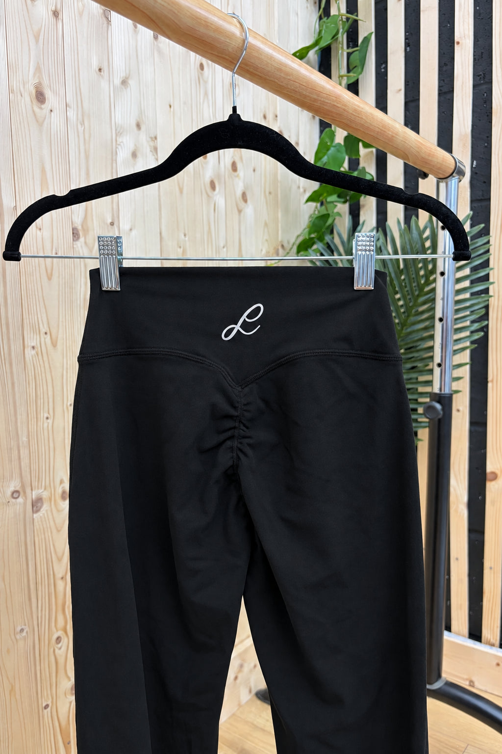 Black high-waisted activewear leggings hanging on a black hanger against a light wood panel wall, featuring a small white embroidered script logo on the back waistband.