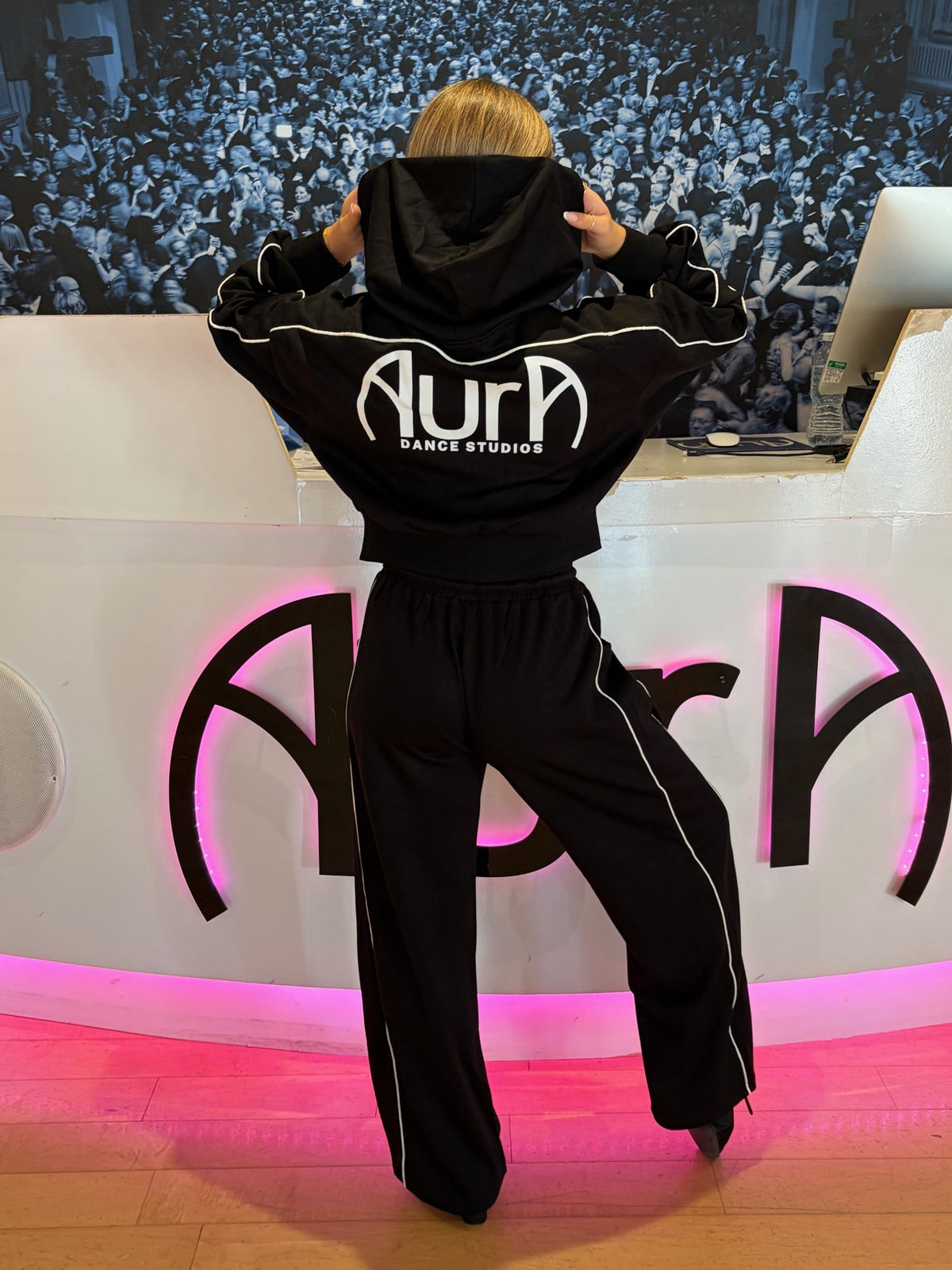 A person stands with their back to the camera wearing a black hoodie and matching sweatpants with white piping and a large white 'Aura DANCE STUDIOS' logo on the back. The setting appears to be a dance studio or event space with a DJ booth, crowd backdrop, and pink illuminated signage.