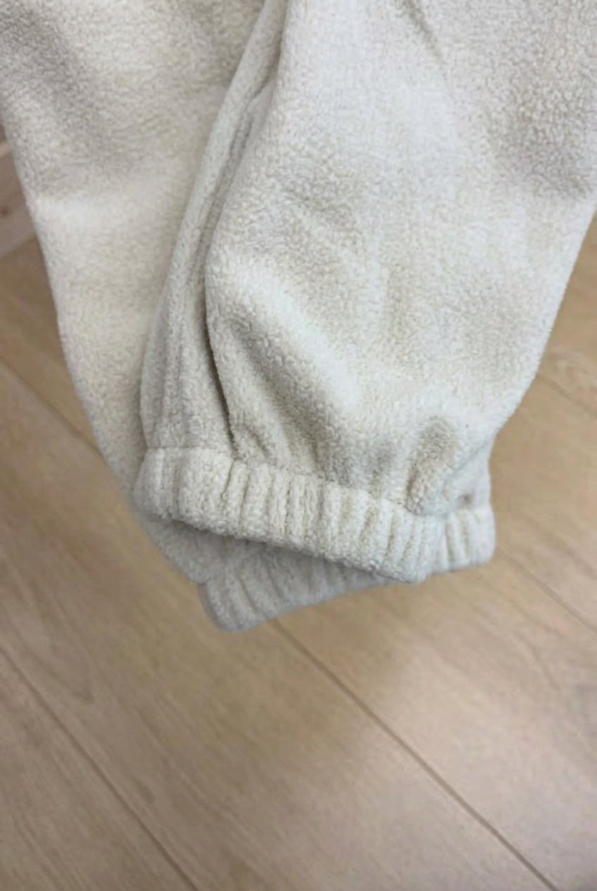 Close-up of a person's hand pulling down the elastic cuff of off-white fleece pants on a light wood floor.