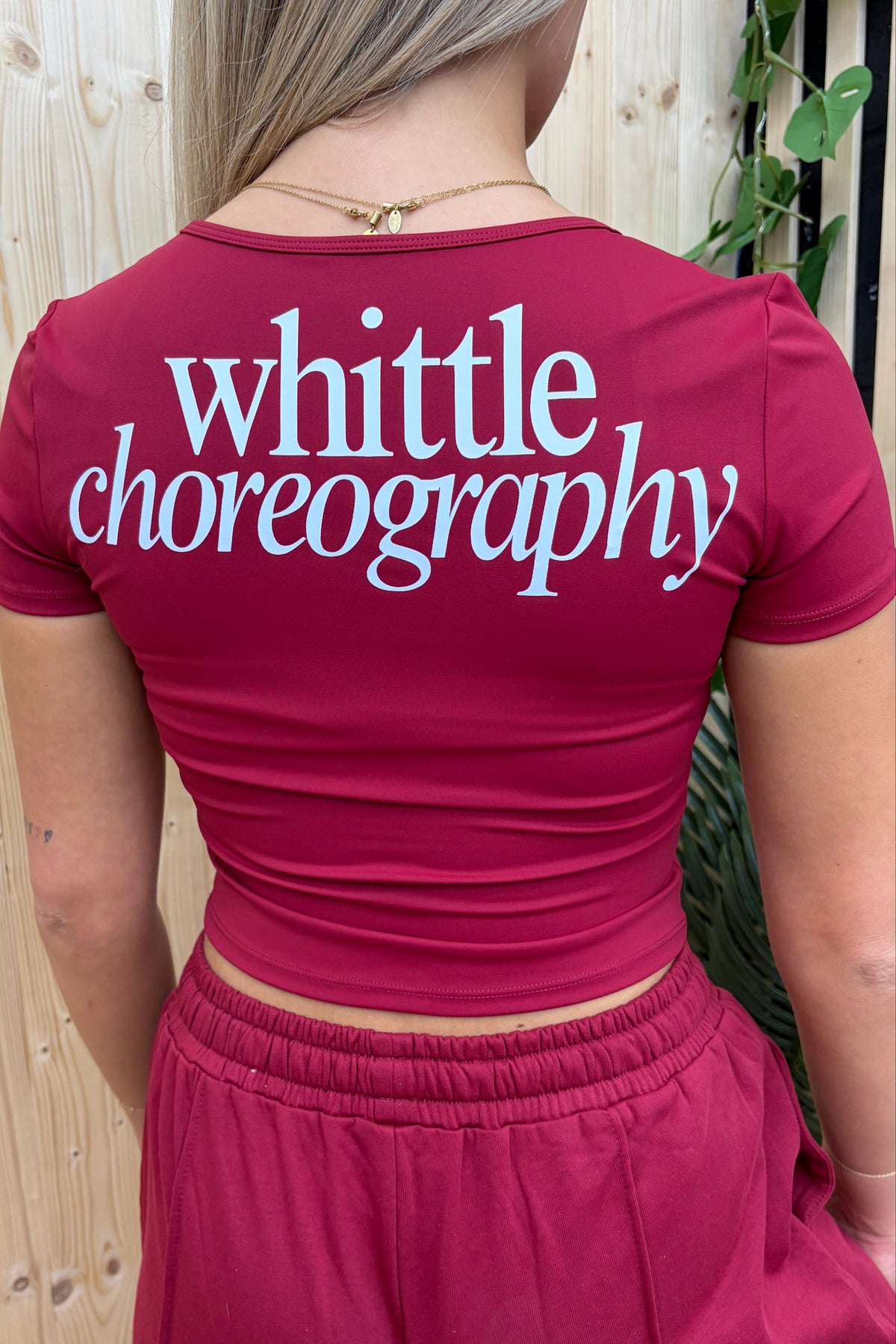 A woman viewed from behind wearing a fitted magenta crop top with the words 'whittle choreography' printed in large white script across the back, paired with matching magenta bottoms.