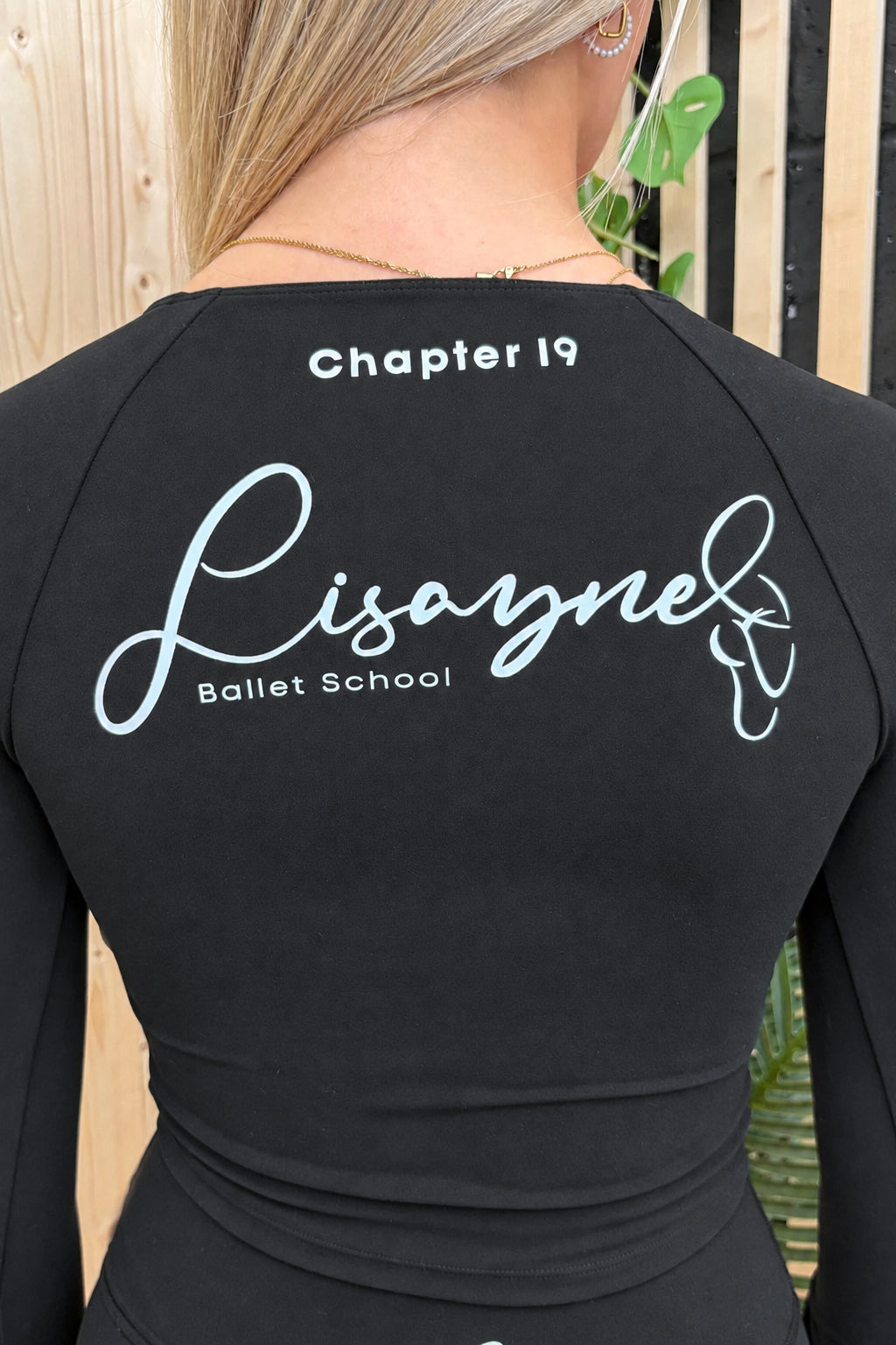Back view of a woman wearing a navy long-sleeve top with 'Chapter 19 Lisaynel Ballet School' printed in white on the back, photographed outdoors against a wooden fence and plants.