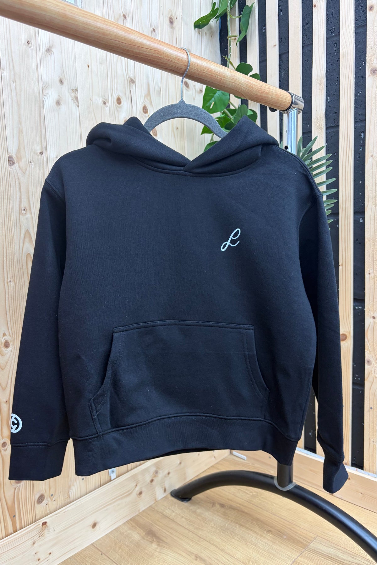 A black pullover hoodie with a small white embroidered script logo on the left chest and a matching logo on the right sleeve, hanging on a black hanger from a wooden clothing rack in front of a light wood slat wall.