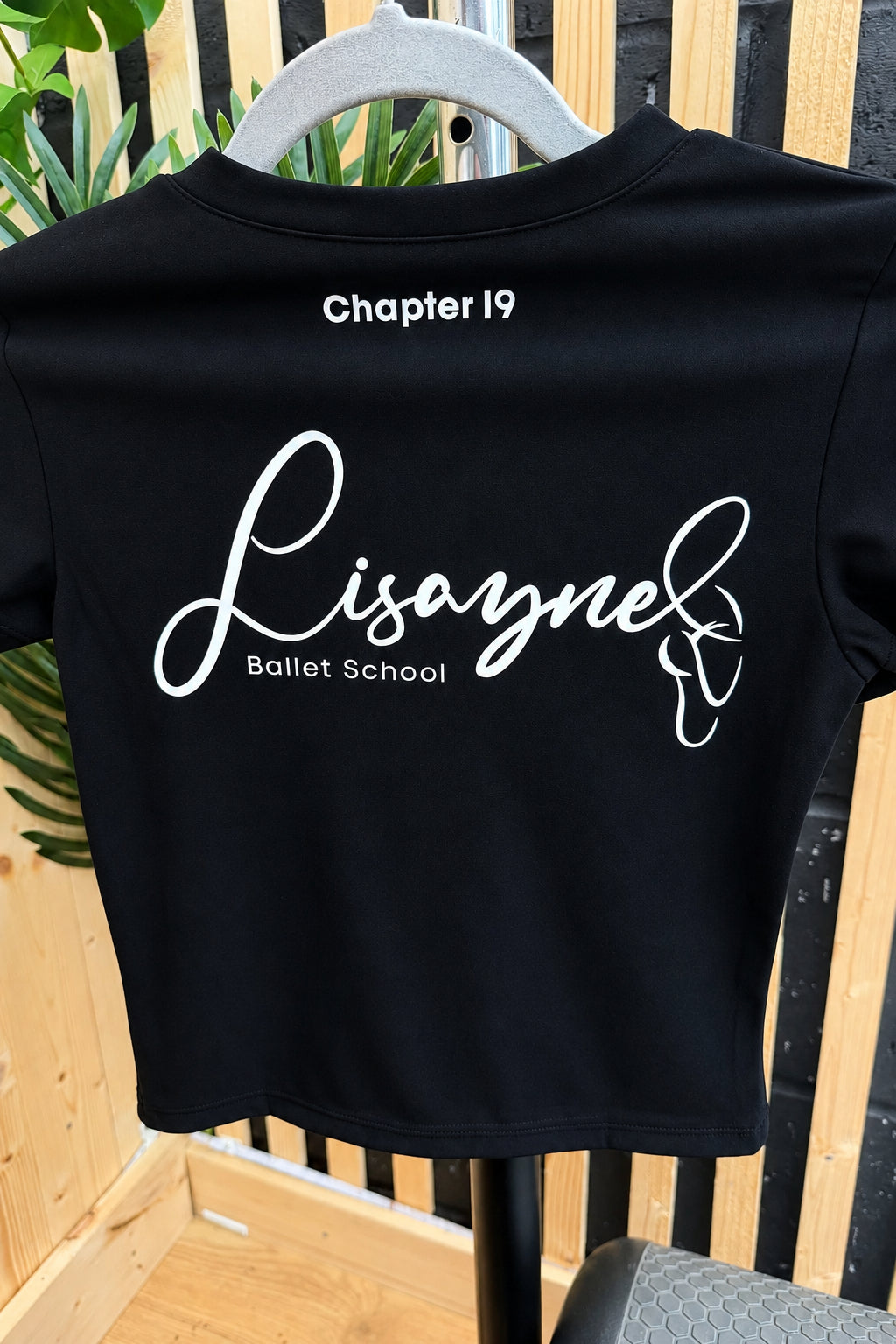 A short-sleeve t-shirt on a hanger, showing the back with white printed text reading 'Chapter 19 Lisayne Ballet School' and a butterfly graphic.