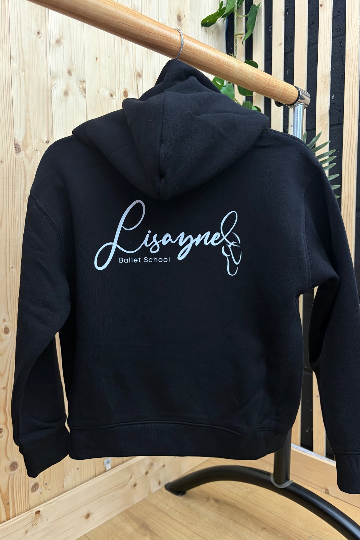 A black hoodie hanging on a black clothing rack, featuring a white script logo on the chest and a circular 'C' logo on the left sleeve cuff. The background consists of light wood paneling and flooring.