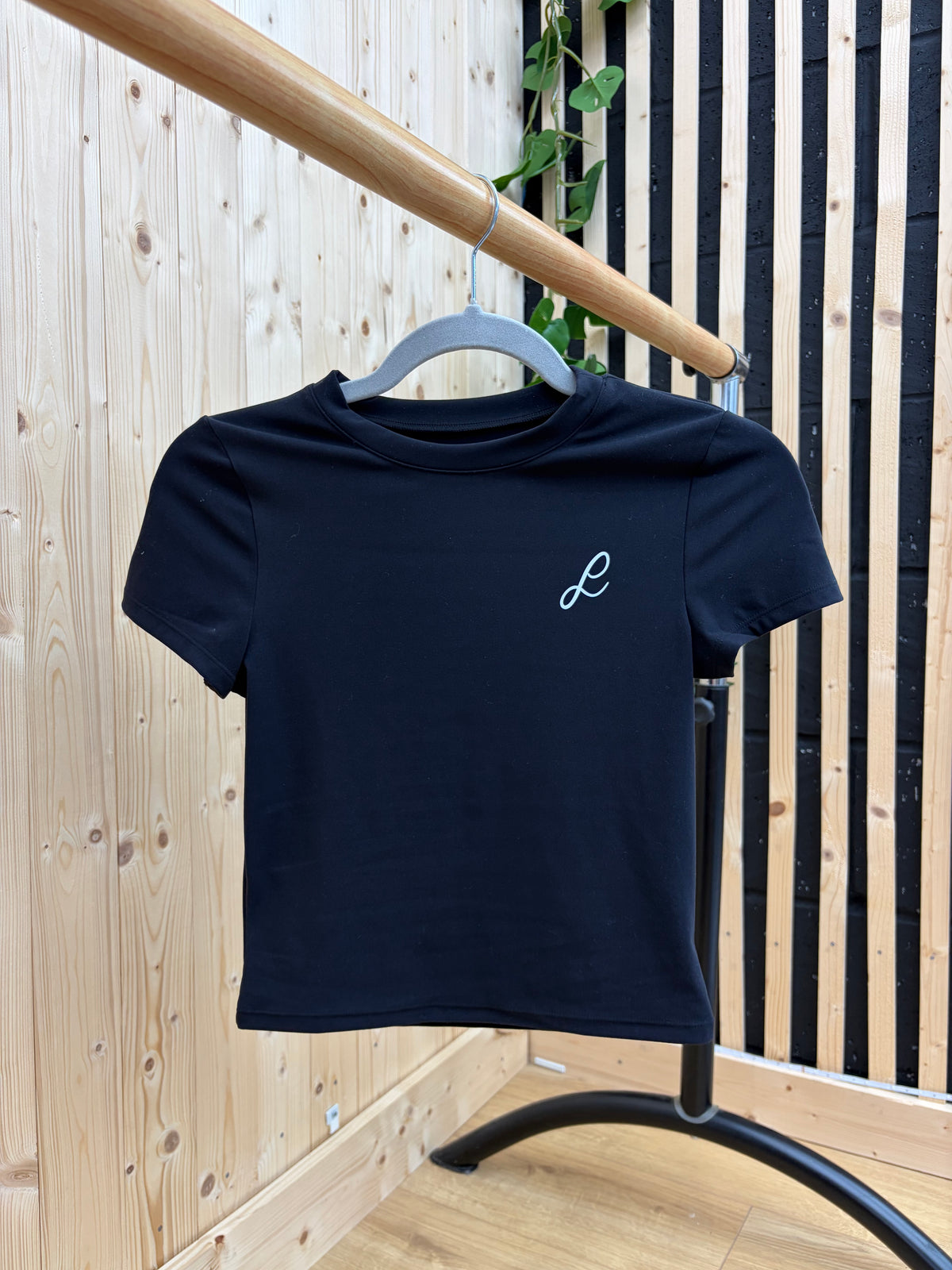 A black short-sleeve crewneck t-shirt with a small white embroidered script 'L' on the left chest, hanging on a gray hanger from a wooden clothing rack in front of a light wood panel wall.
