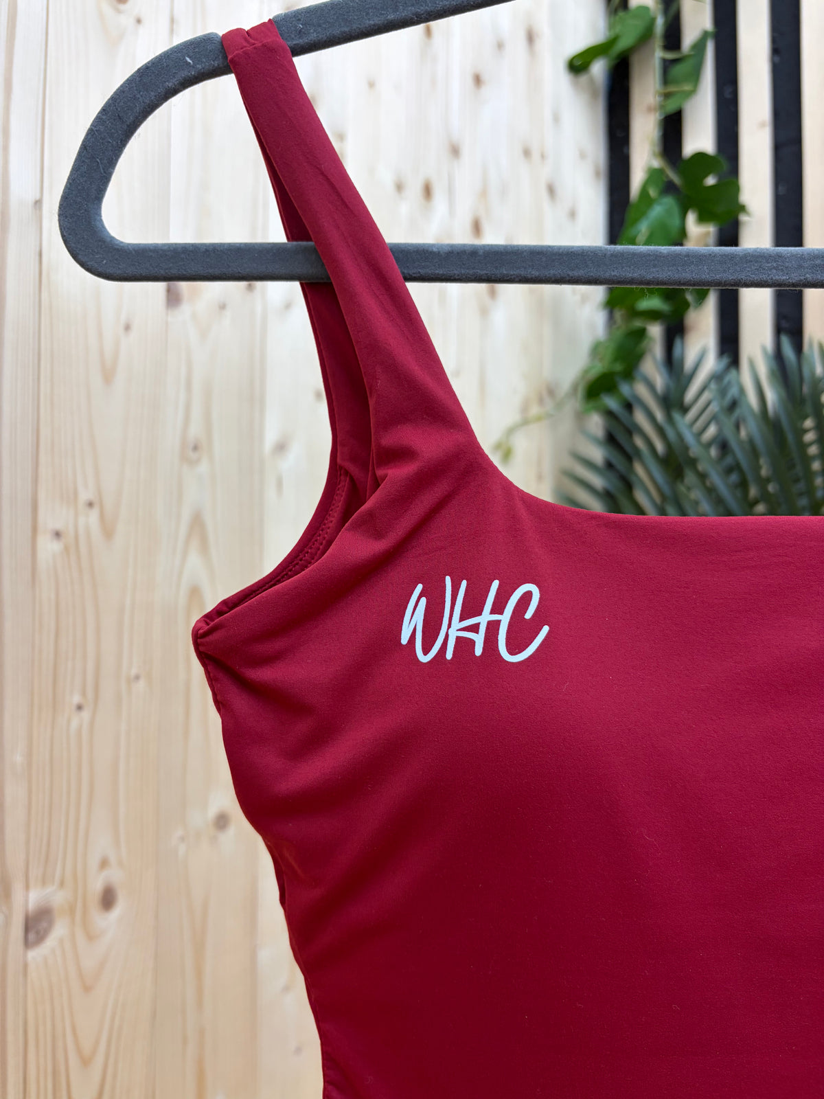 A close-up of a dark red tank top hanging on a black metal hanger against a light wood panel wall, featuring the white embroidered initials 'WHC' on the chest.