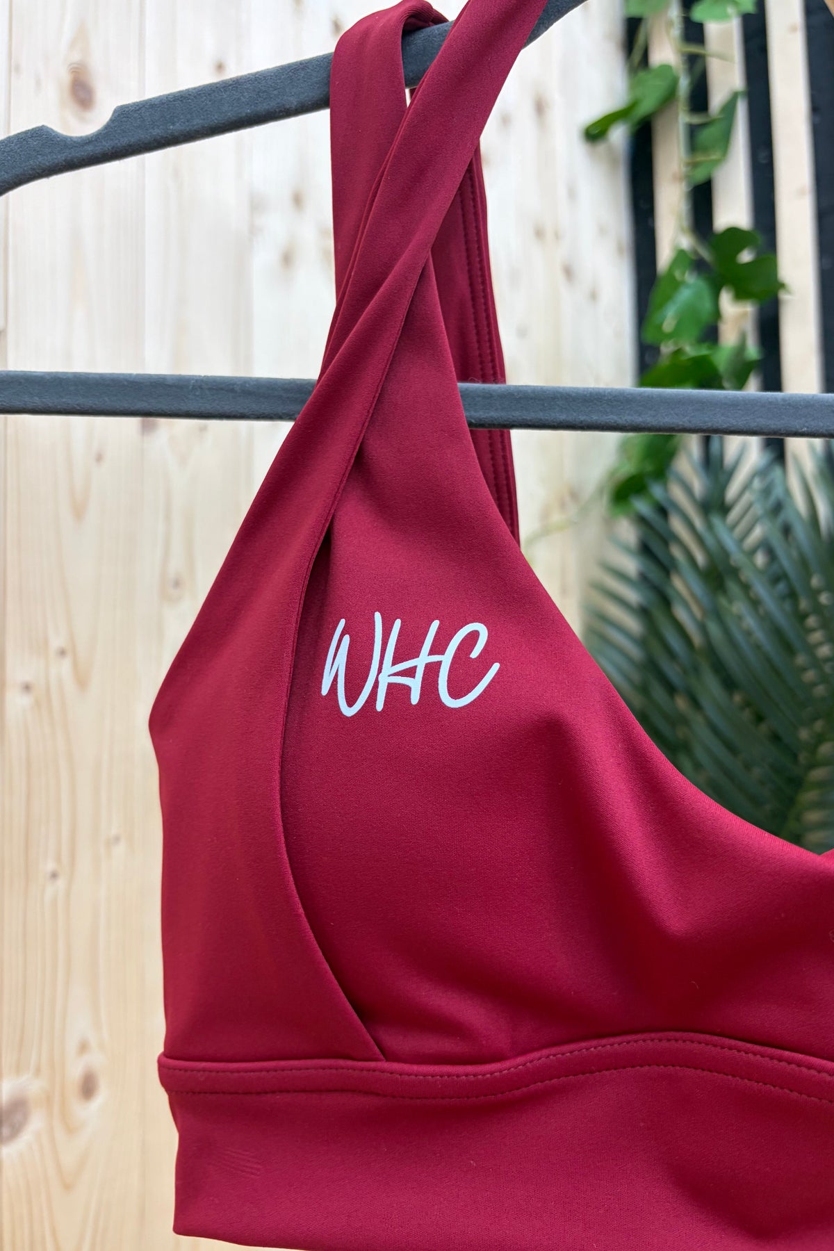 A close-up of a dark red sports bra hanging on a black hanger against a light wood panel wall, with green leaves in the background. The word 'WHC' is printed in white script on the front.