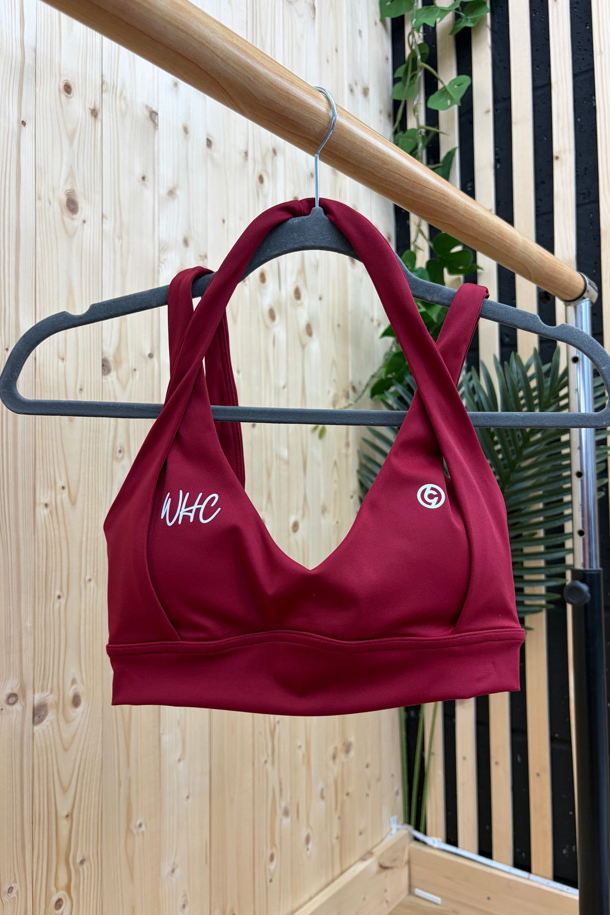 A burgundy sports bra with white embroidered logos hanging on a black hanger from a wooden clothes rack, set against a light wood panel wall with green plants.