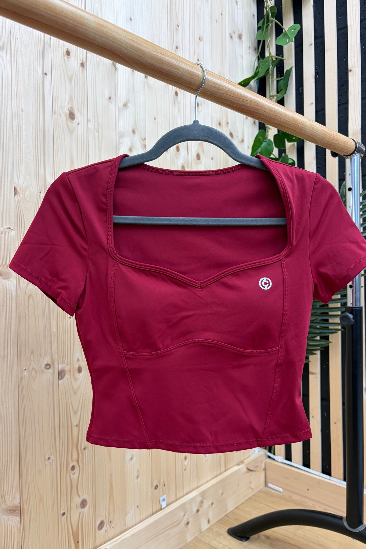 A maroon short-sleeve crop top with a square neckline and front seam detailing, hanging on a black hanger from a wooden clothing rack against a light wood panel wall.