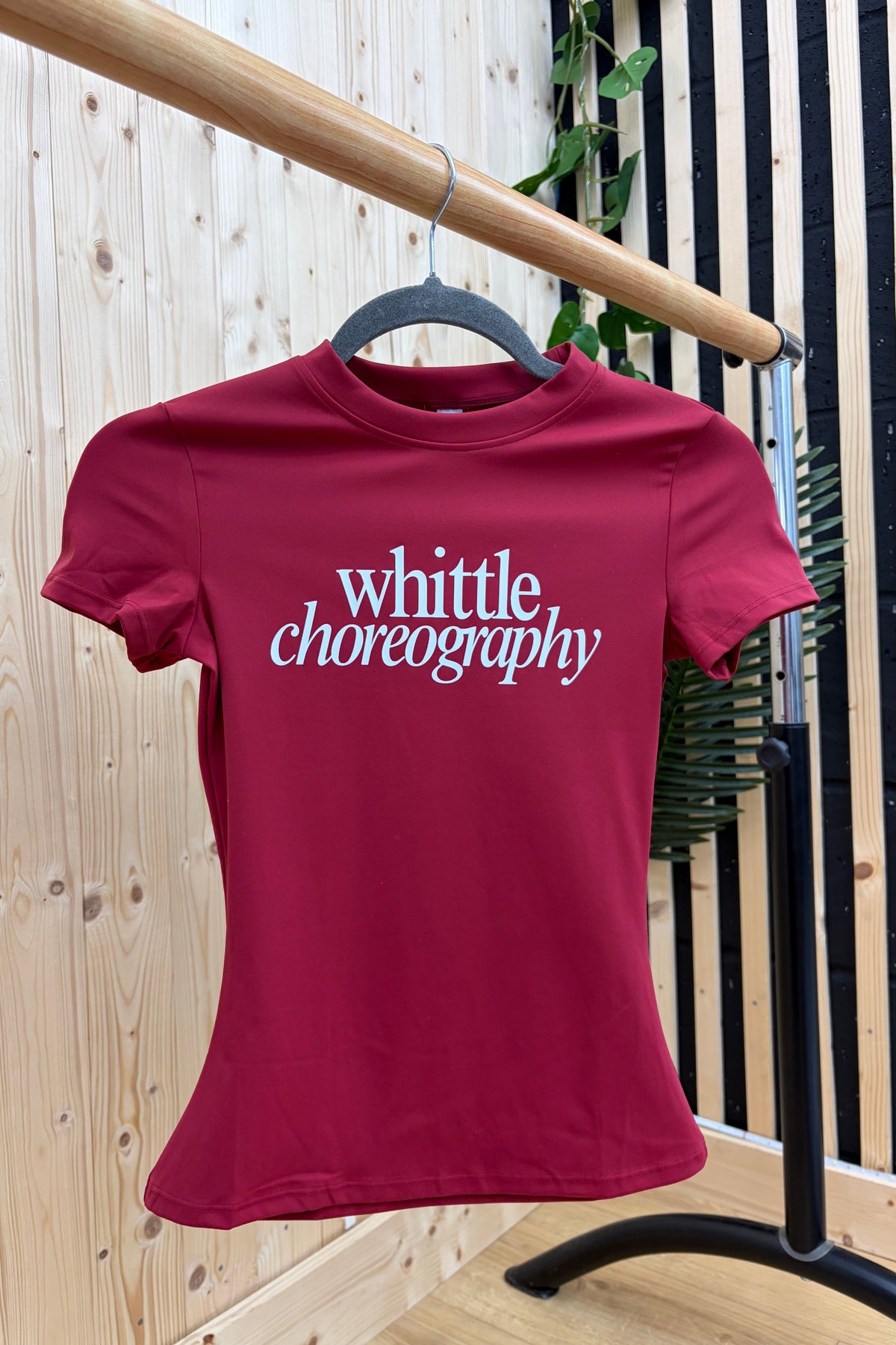 A fitted short-sleeve crewneck t-shirt in deep burgundy red, hanging on a black hanger from a wooden clothing rack. The shirt features the words 'whittle choreography' printed in white script across the chest.