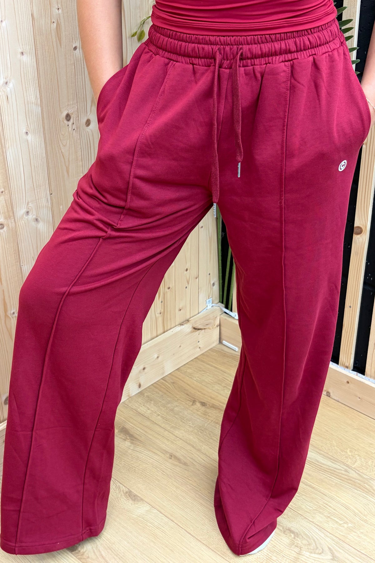 A person wearing wide-leg sweatpants in a deep berry pink or burgundy, standing indoors against a light wood plank wall and floor. The photo is cropped from the waist down, showing the pants' fit, drawstring waist, side pockets, and a small white logo on the upper right thigh.