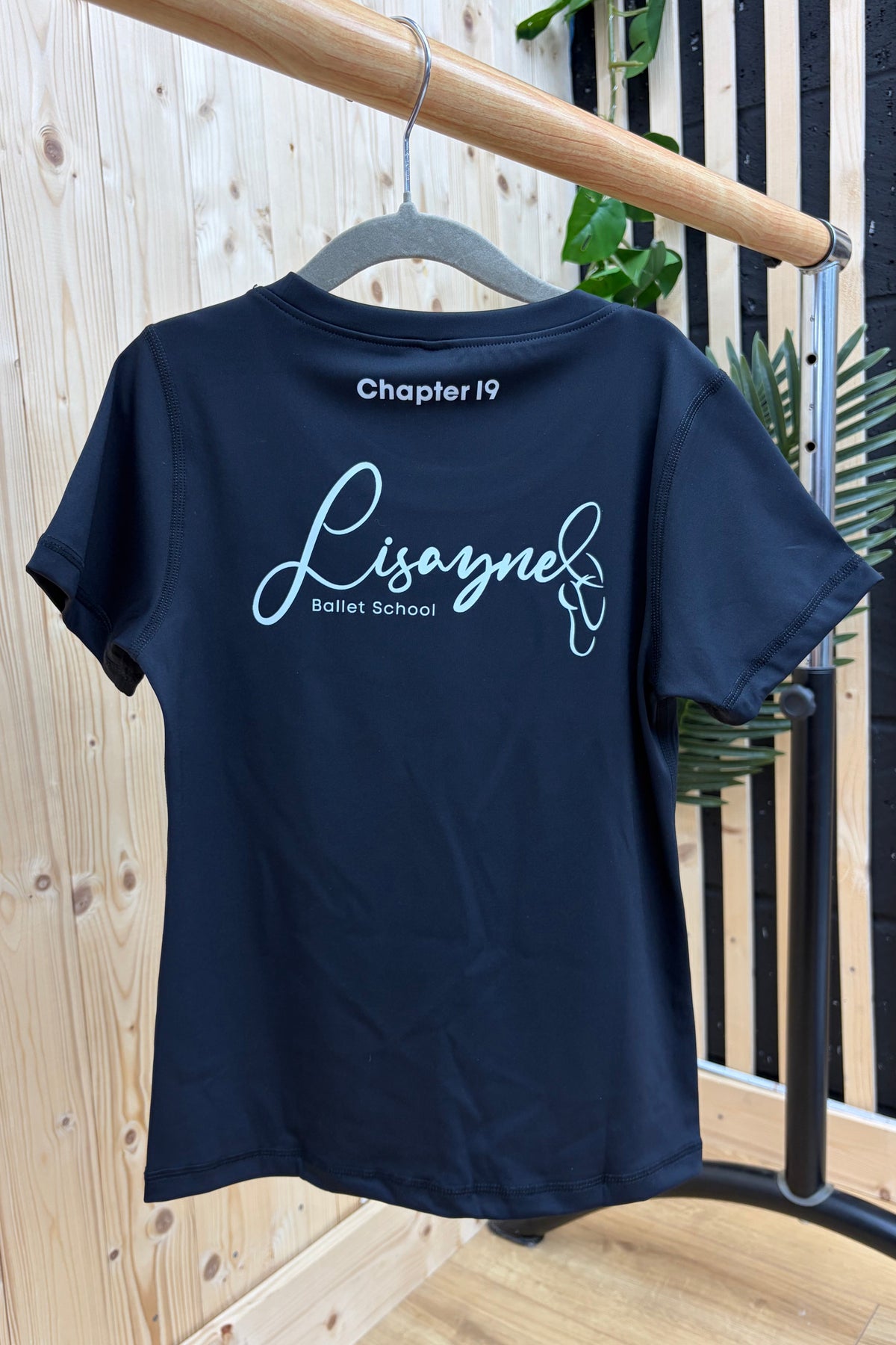 A short-sleeve t-shirt hanging on a gray hanger from a wooden clothing rack. The back of the shirt features a white graphic print reading 'Chapter 19 Lisaynel Bismischool'.