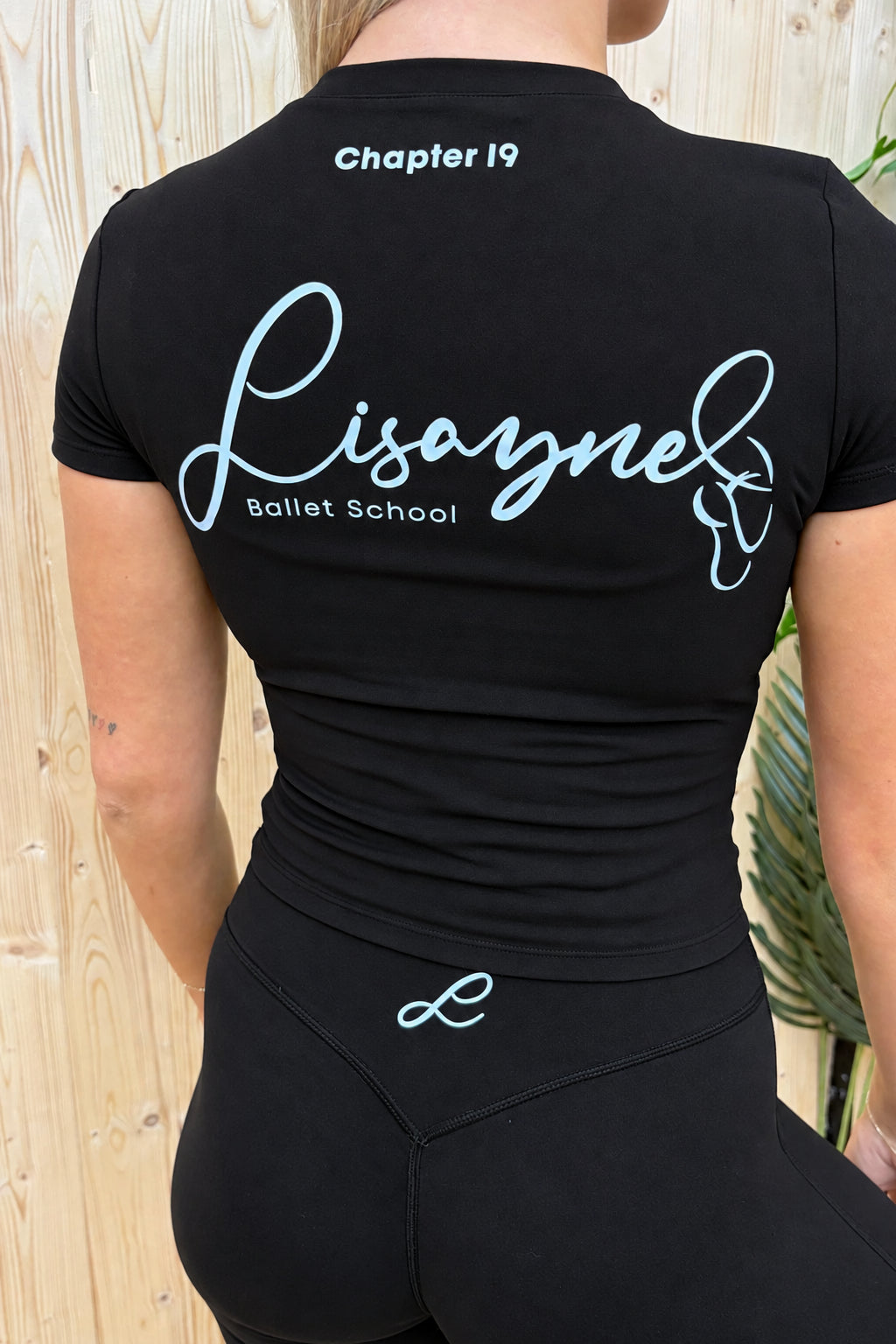 A woman shown from the back wearing a navy blue fitted athletic top and matching leggings. The top has white script text reading 'Lisayne Ballet School' and 'Chapter 19' near the collar, with a small white script logo near the waistband.