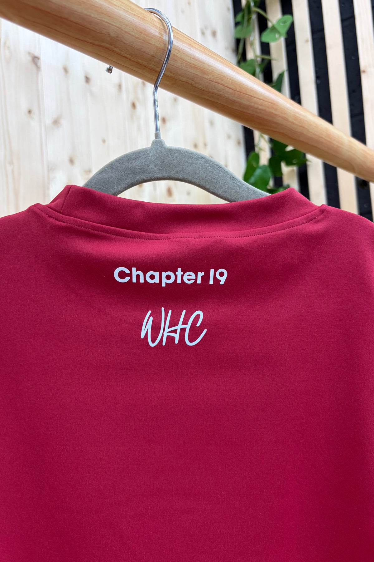 A close-up of a dark red crewneck sweatshirt hanging on a gray hanger from a wooden clothes rack. The back of the sweatshirt features white printed text reading 'Chapter 19' and 'WHC'.