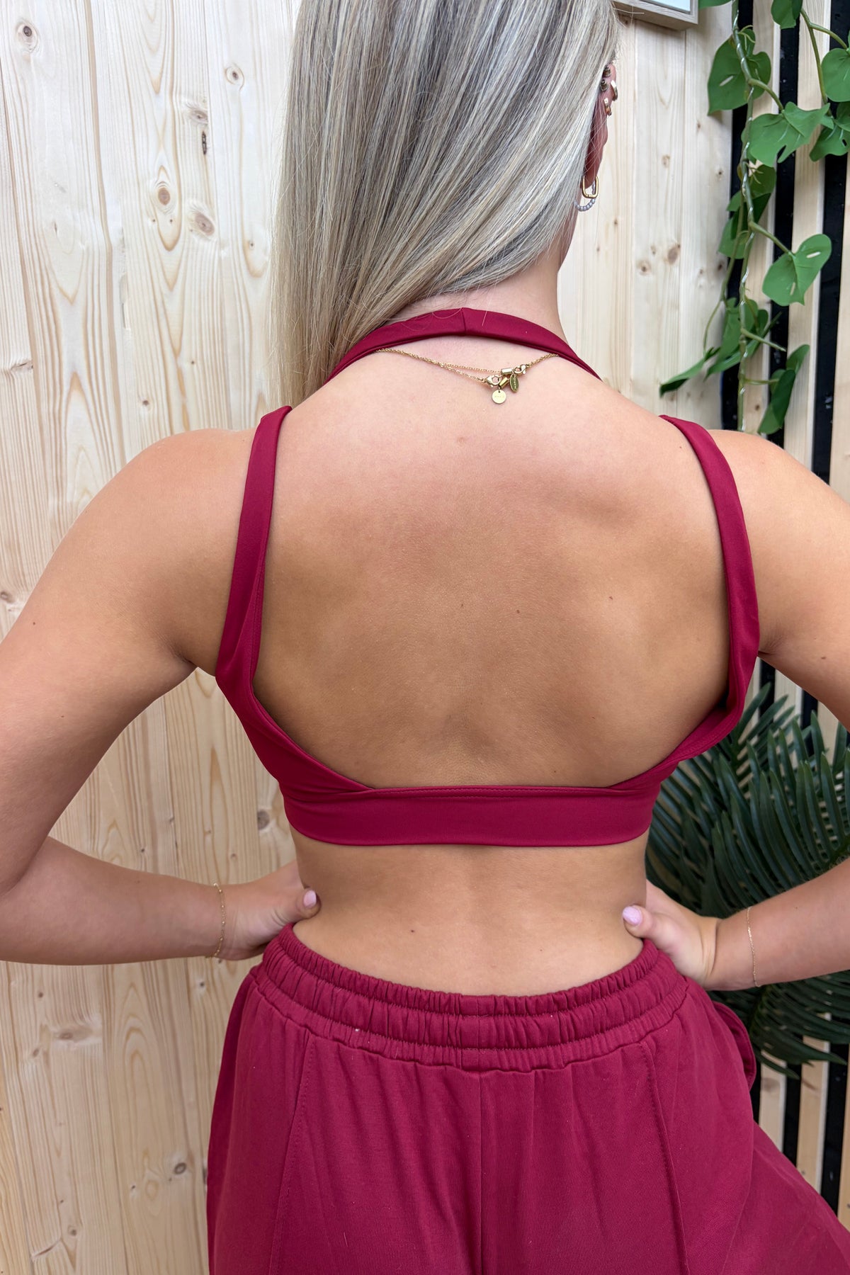 A dark red sports bra with a racerback design hanging on a gray hanger from a wooden clothes rack, set against a light wood panel wall with a black and white striped screen and green plants.