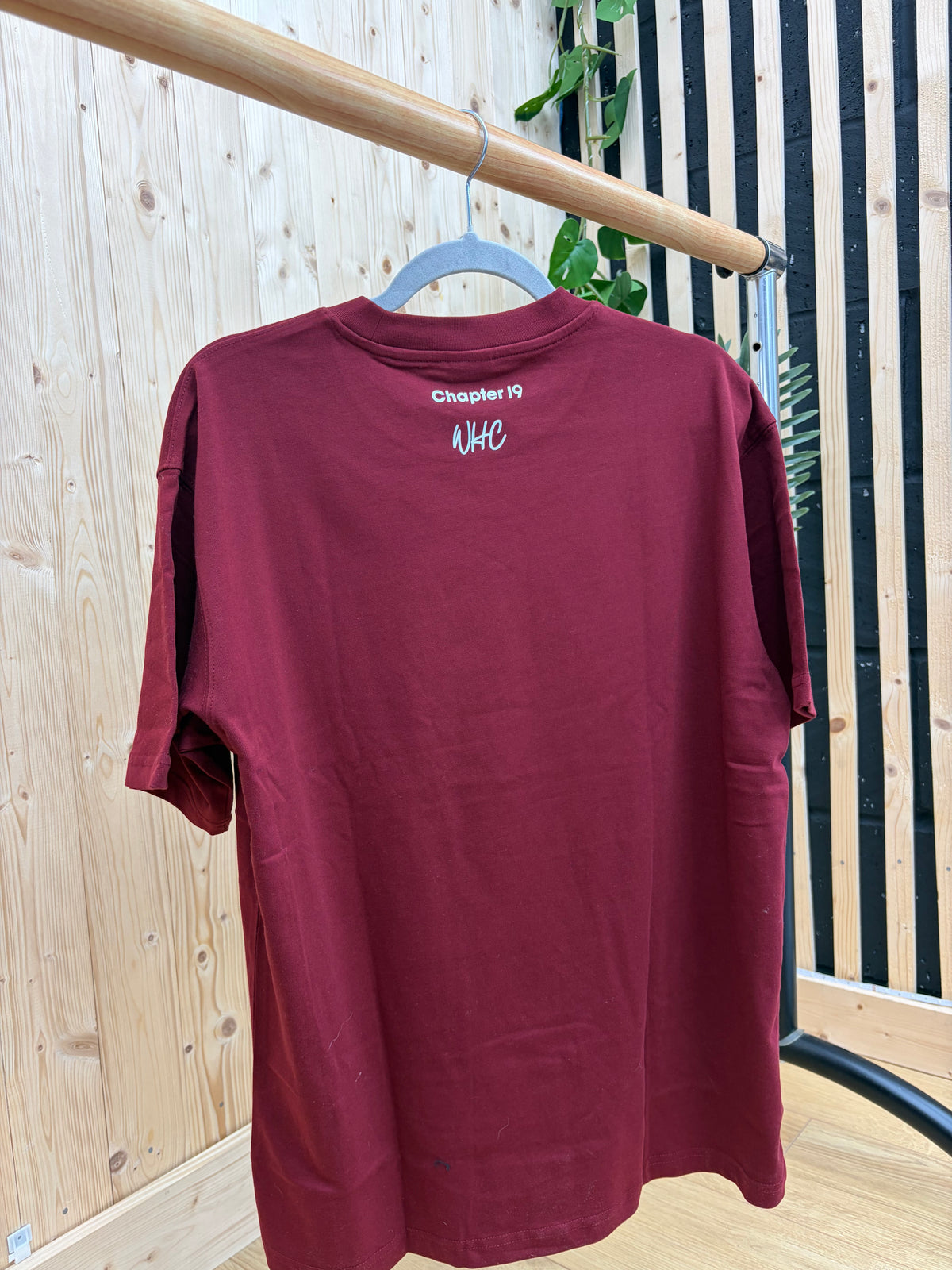 A maroon short-sleeve t-shirt hanging on a blue hanger from a wooden clothing rack, positioned against a background of light wood paneling and black-and-white vertical slats.