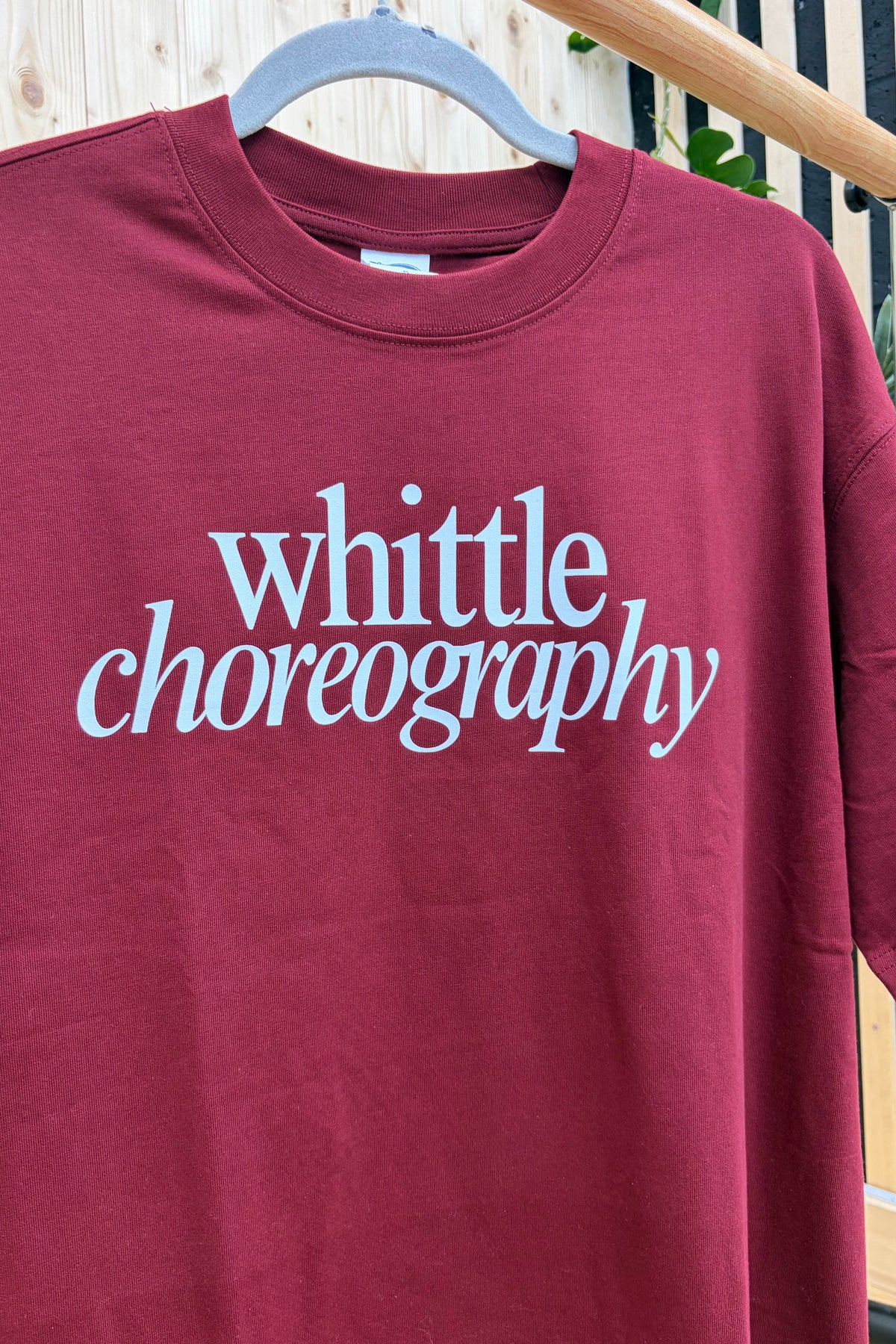 A maroon crewneck t-shirt hanging on a wooden hanger, featuring the words 'whittle choreography' printed in a light blue cursive font across the chest.