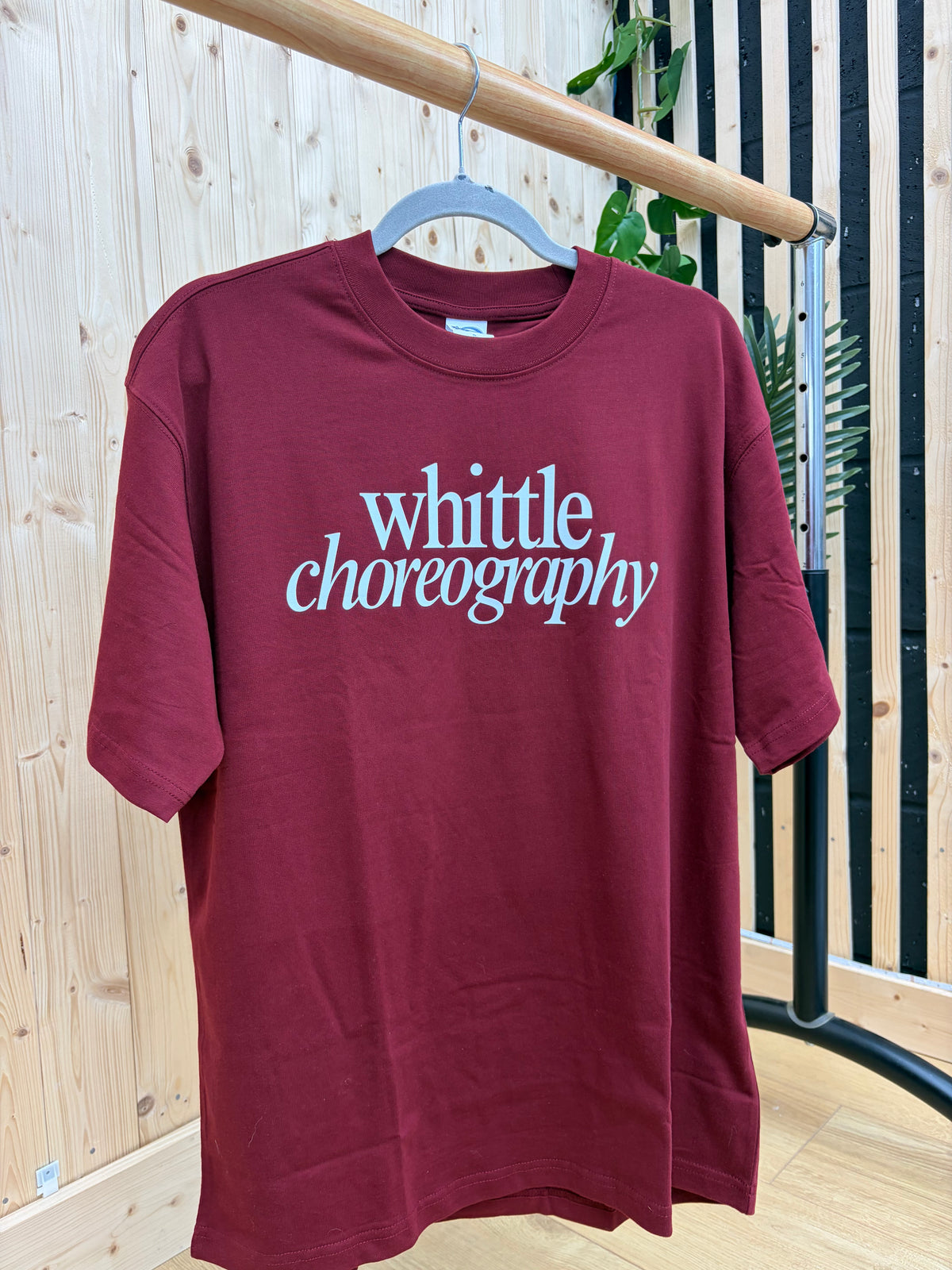 A maroon short-sleeve t-shirt hanging on a hanger from a wooden clothing rack. The shirt features the words 'whittle choreography' printed in a stylized white font across the chest.
