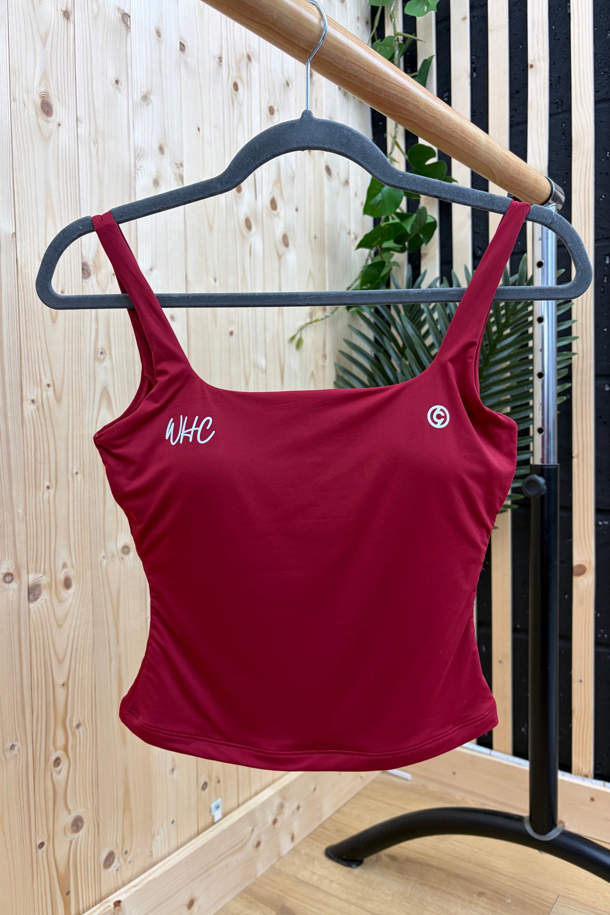 A maroon athletic crop top with white embroidered 'NHC' lettering on the left chest and a small circular logo on the right chest, hanging on a black hanger on a clothing rack against a light wood panel wall.