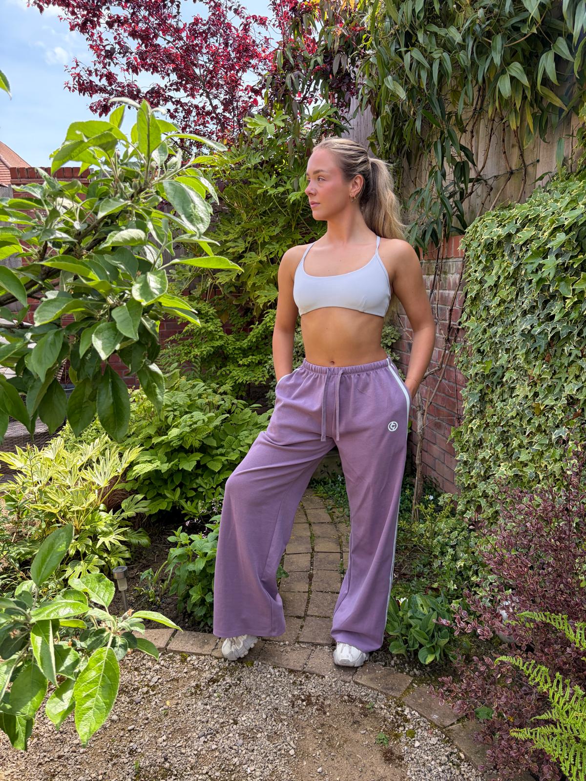 Woman in a garden wearing a light blue sports bra and purple sweatpants.